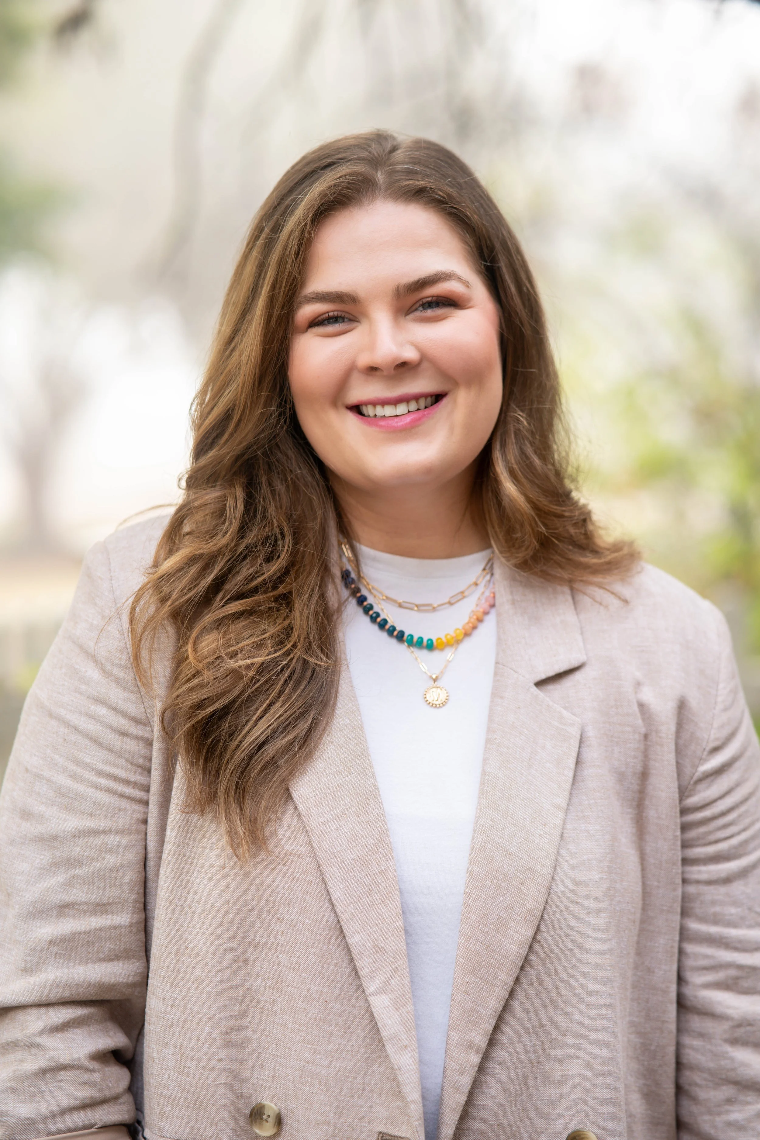 Close-up of a woman with long brown wavy hair, wearing a beige blazer and layered necklaces, smiling outdoors with a blurred tree background.