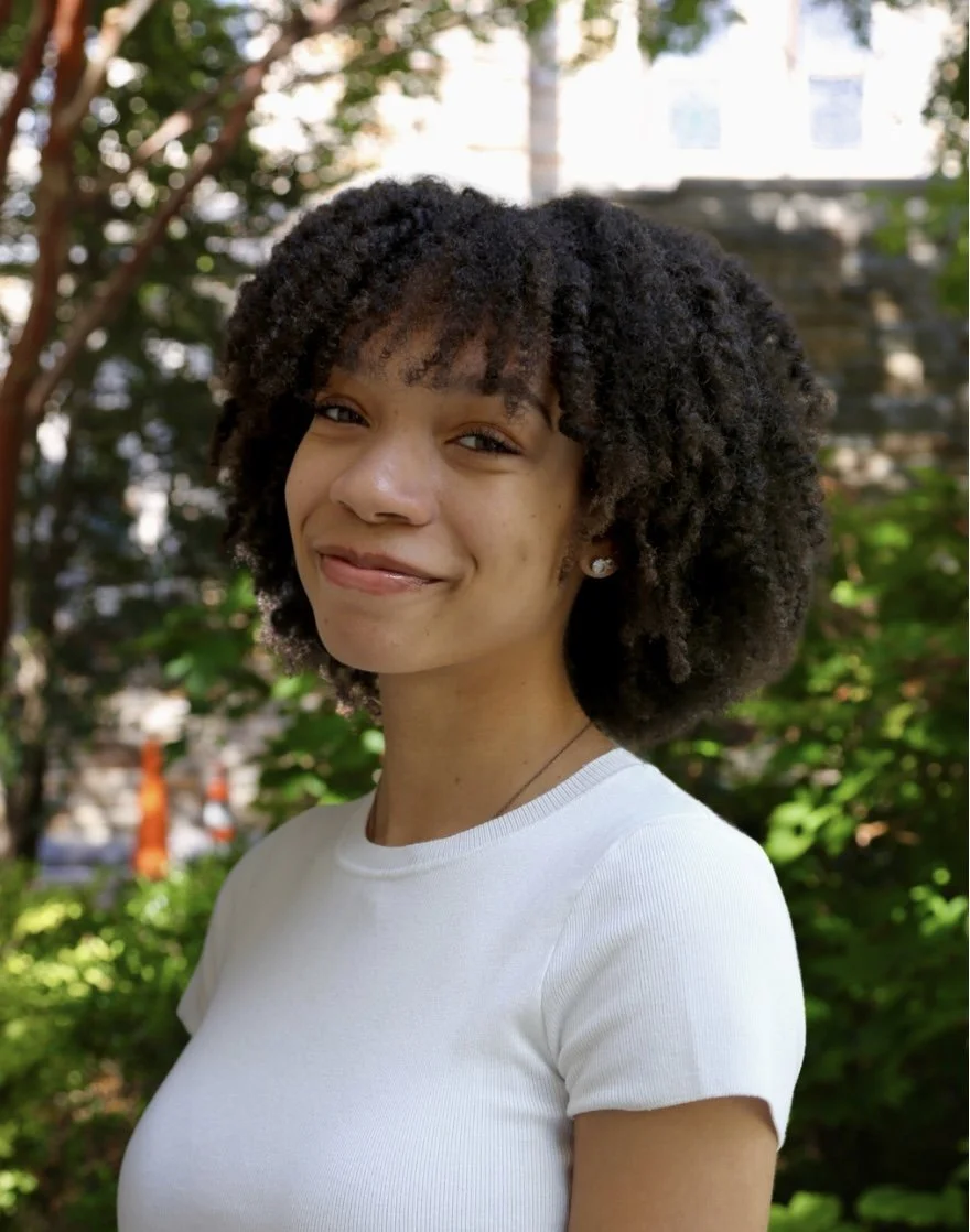 A young woman with natural curly hair smiling outdoors, wearing a white shirt and small earrings, with trees and a building in the background.