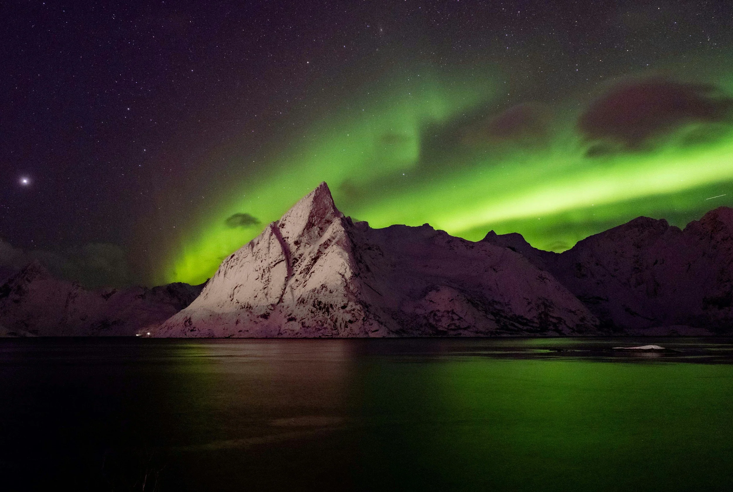 Nighttime scene of snow-covered mountain with green northern lights in the starry sky above a body of water.