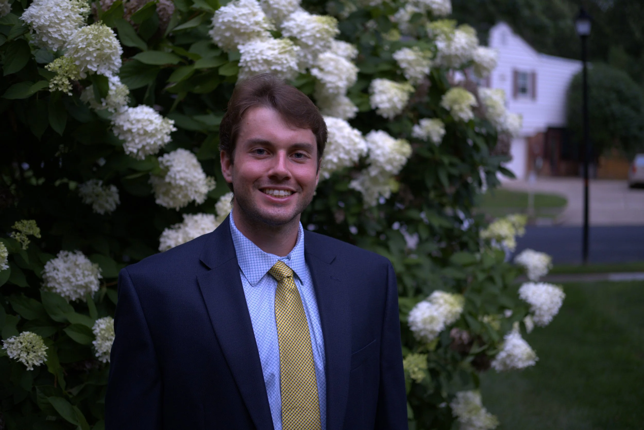 A young man wearing a navy blazer and a yellow tie standing in front of a bush with white flowers.