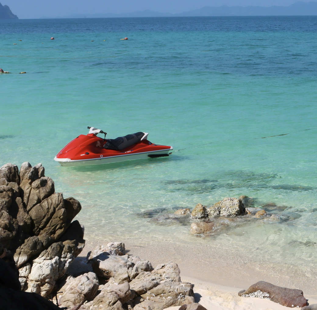 A red jet ski floating near the shoreline on clear, turquoise water with rocks and a sandy beach in the foreground and distant mountains on the horizon.