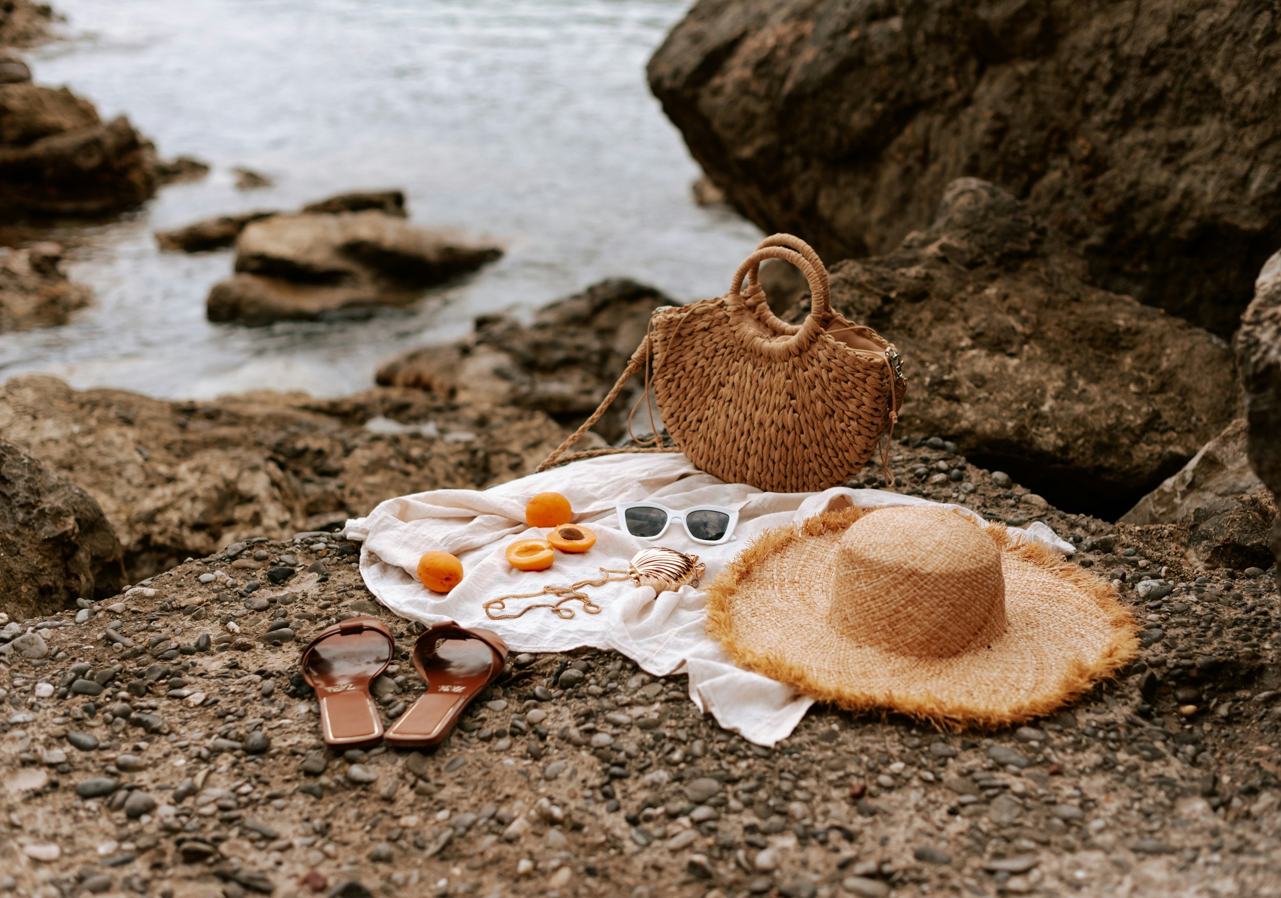 Beach setup with a straw wide-brimmed hat, woven bag, sunglasses, and flip-flops on a white cloth among rocks by the water.