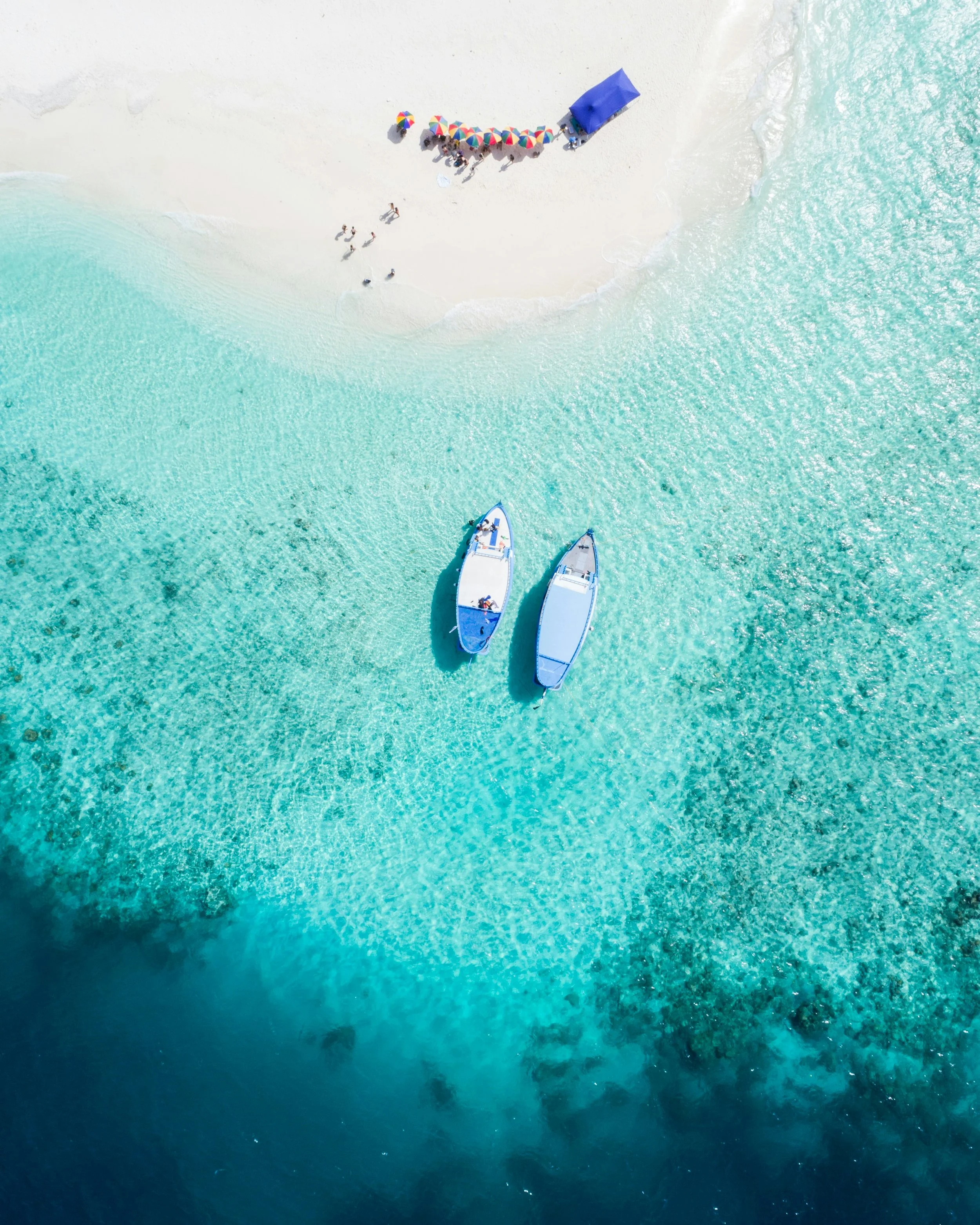 Aerial view of a sandy beach with two boats floating in turquoise water and a group of people under a colorful umbrellas on the shore.
