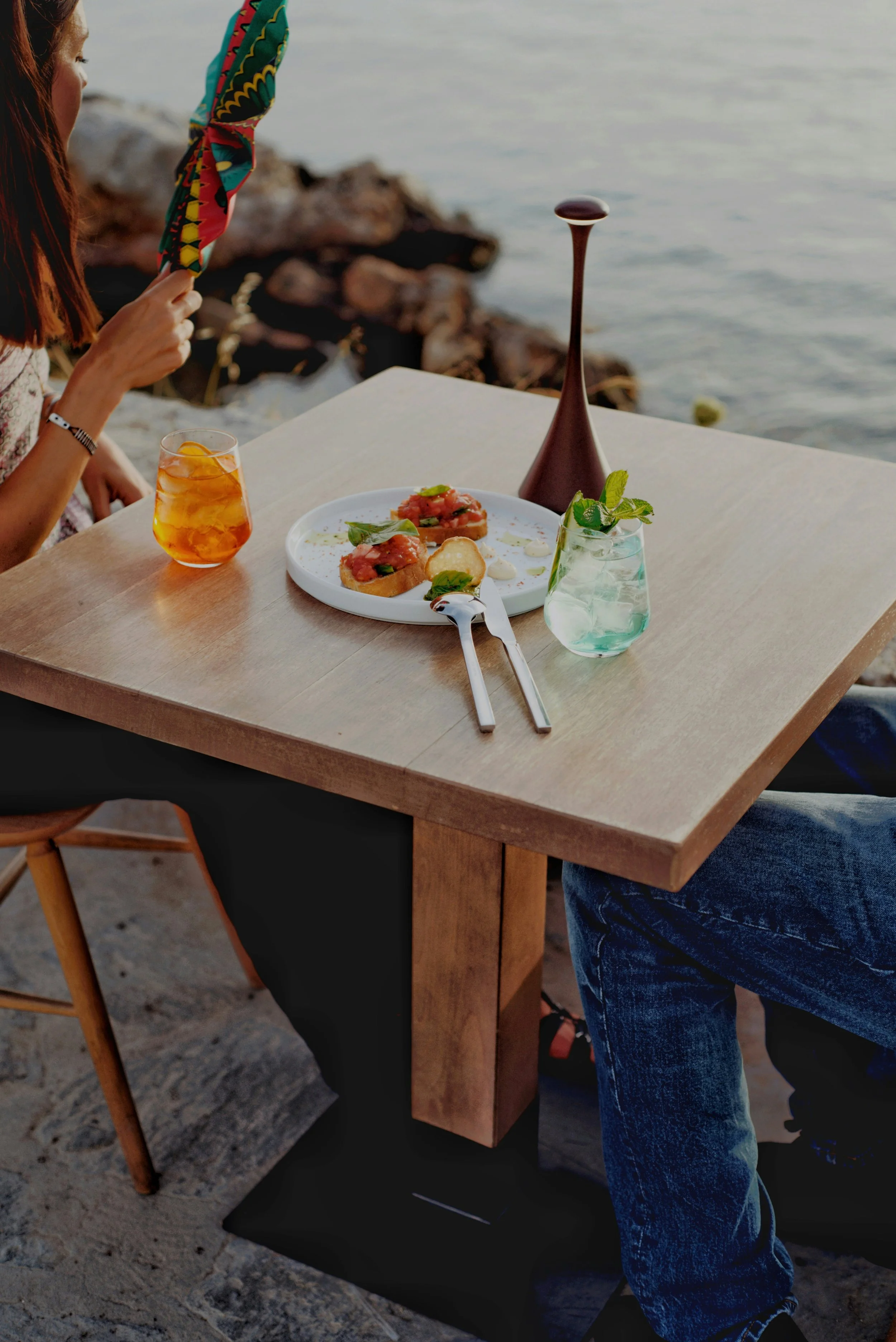 A woman and a man sitting at a wooden outdoor table by the water. The table has drinks and a plate with food, possibly appetizers or small dishes. The woman is holding a colorful scarf and there is a tall, narrow decorative object on the table.