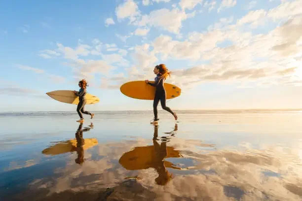 Two women carrying surfboards walk on a wet beach during sunset or sunrise, with reflections on the water and a partly cloudy sky.