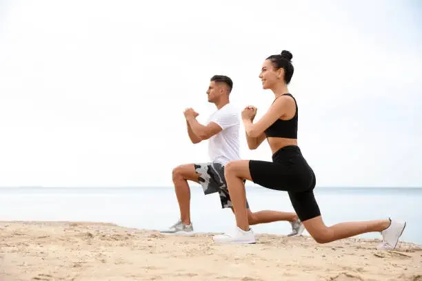 A man and woman exercising on the beach, doing lunges with hands clasped in front of their chests, facing each other.