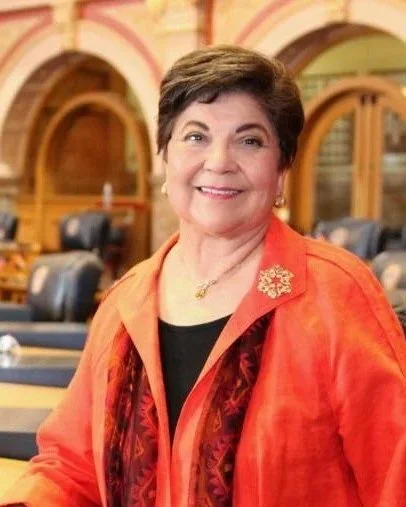 A woman with short brown hair smiling, wearing an orange jacket with a gold brooch, in a formal setting with wooden decor and leather chairs.