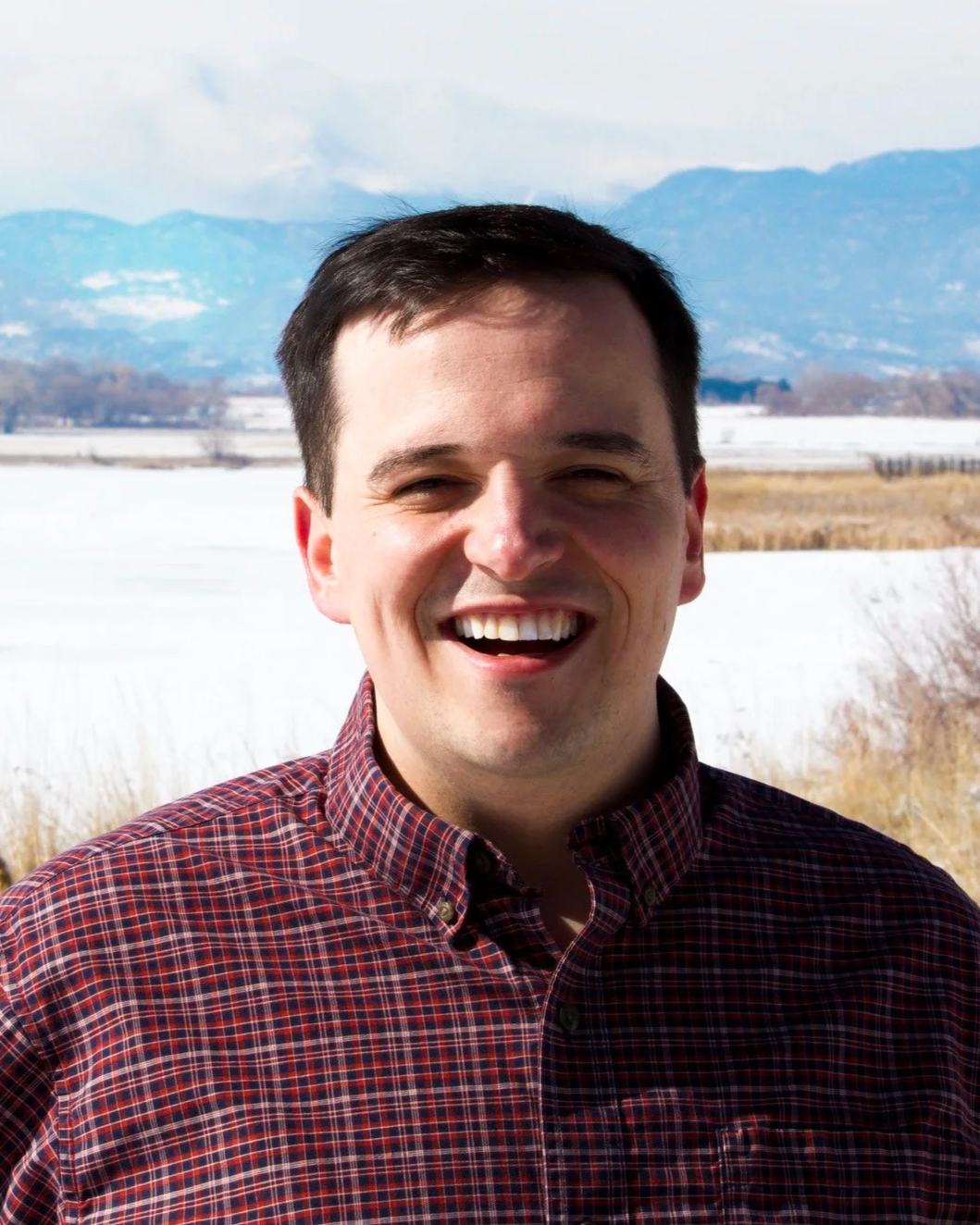 A man smiling outdoors in a winter landscape with snow-covered fields and mountains in the background.