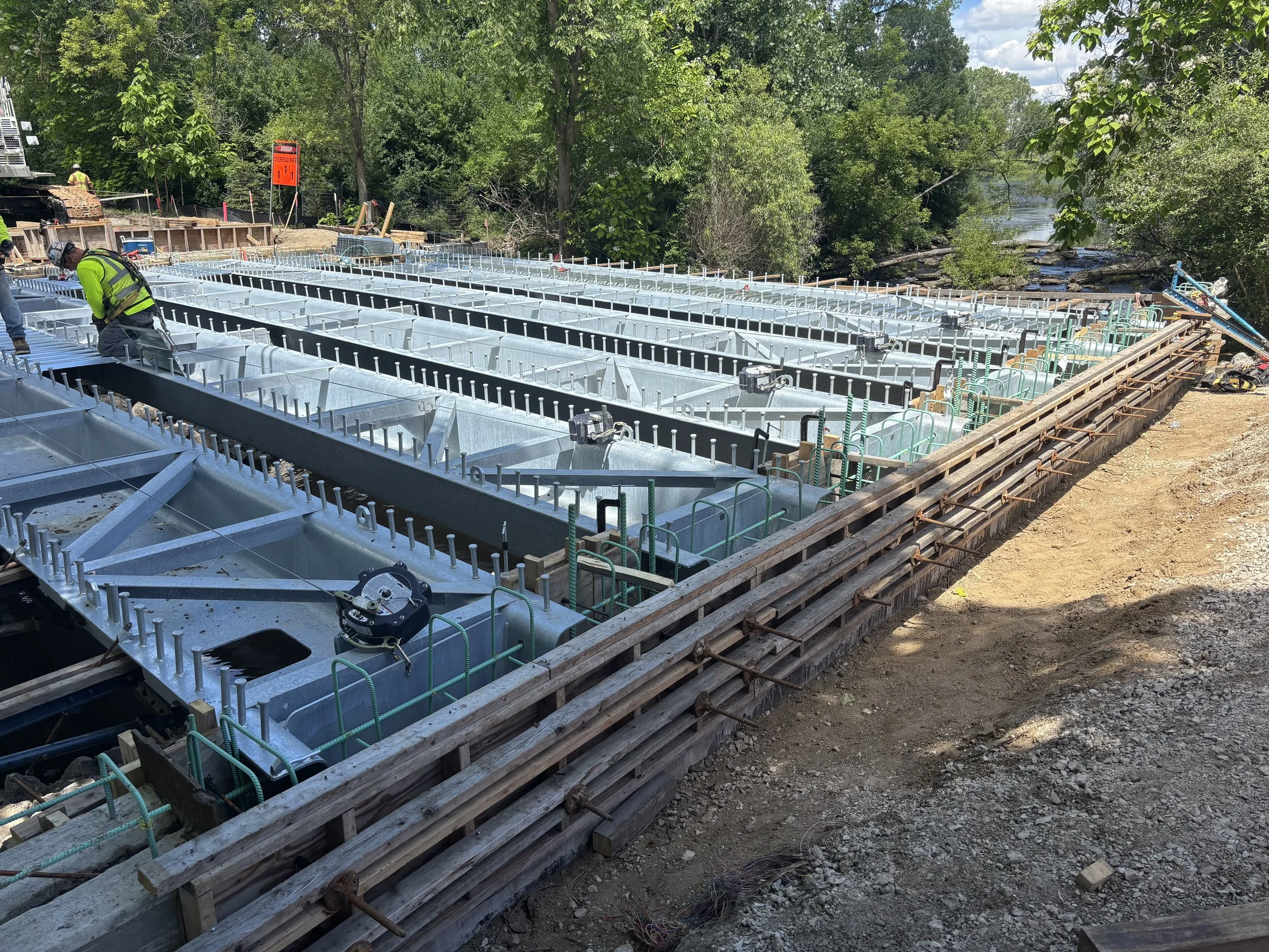 Construction workers building a bridge over a river, with metal framework and wooden formwork in place, surrounded by green trees.