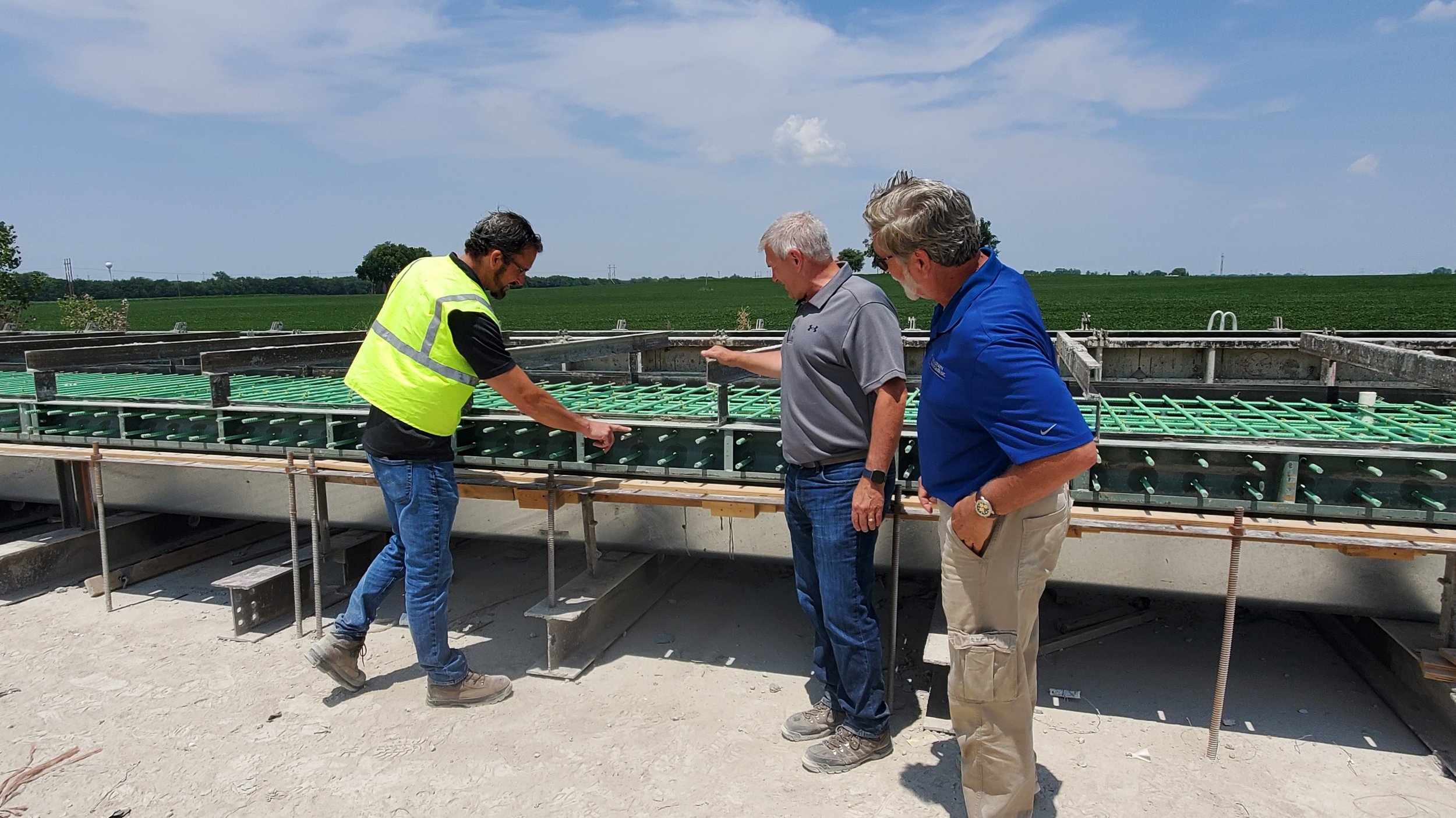 Three men examining a construction site with green rebar in a concrete foundation, with a rural landscape and blue sky in the background.