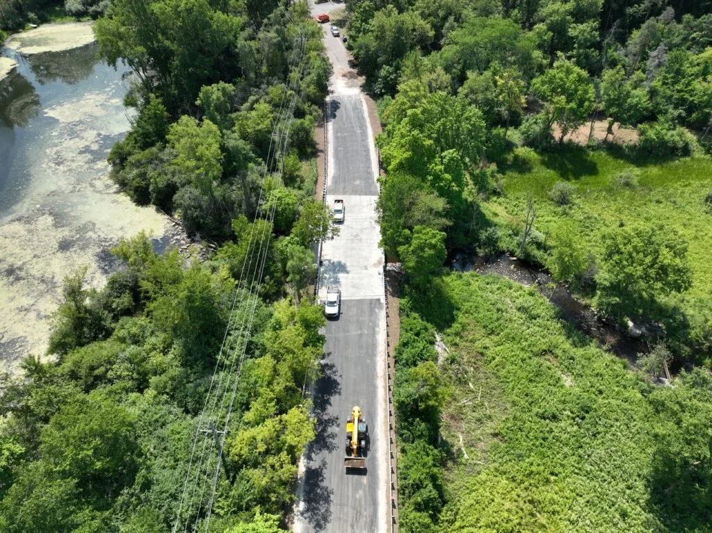 Construction workers building a bridge over a river, with metal framework and wooden formwork in place, surrounded by green trees.