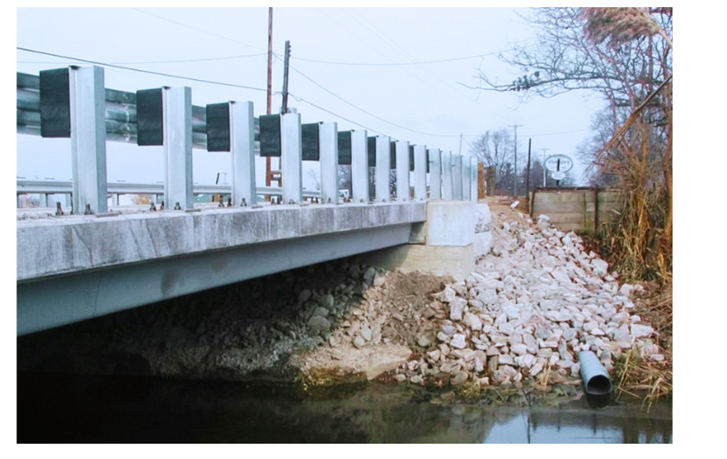 View of a bridge over a waterway with gravel and rocks along the shoreline, and a sewer pipe extending into the water.