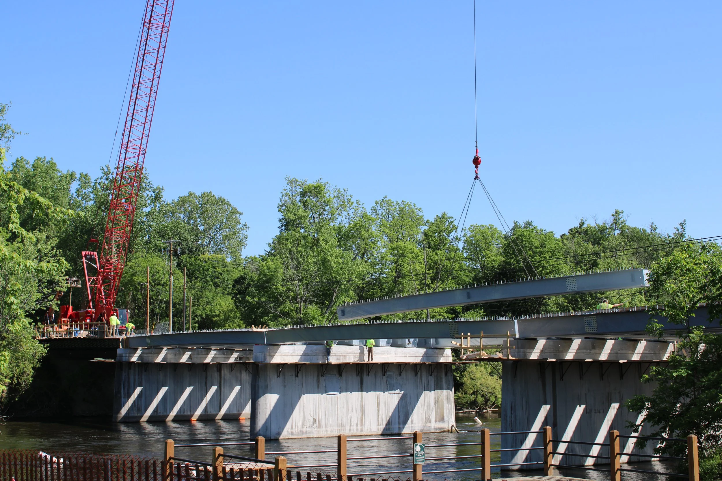 Construction workers standing on a bridge under construction, with a large red crane lifting a section of the bridge into place, greenery in the background, and a bright blue sky.