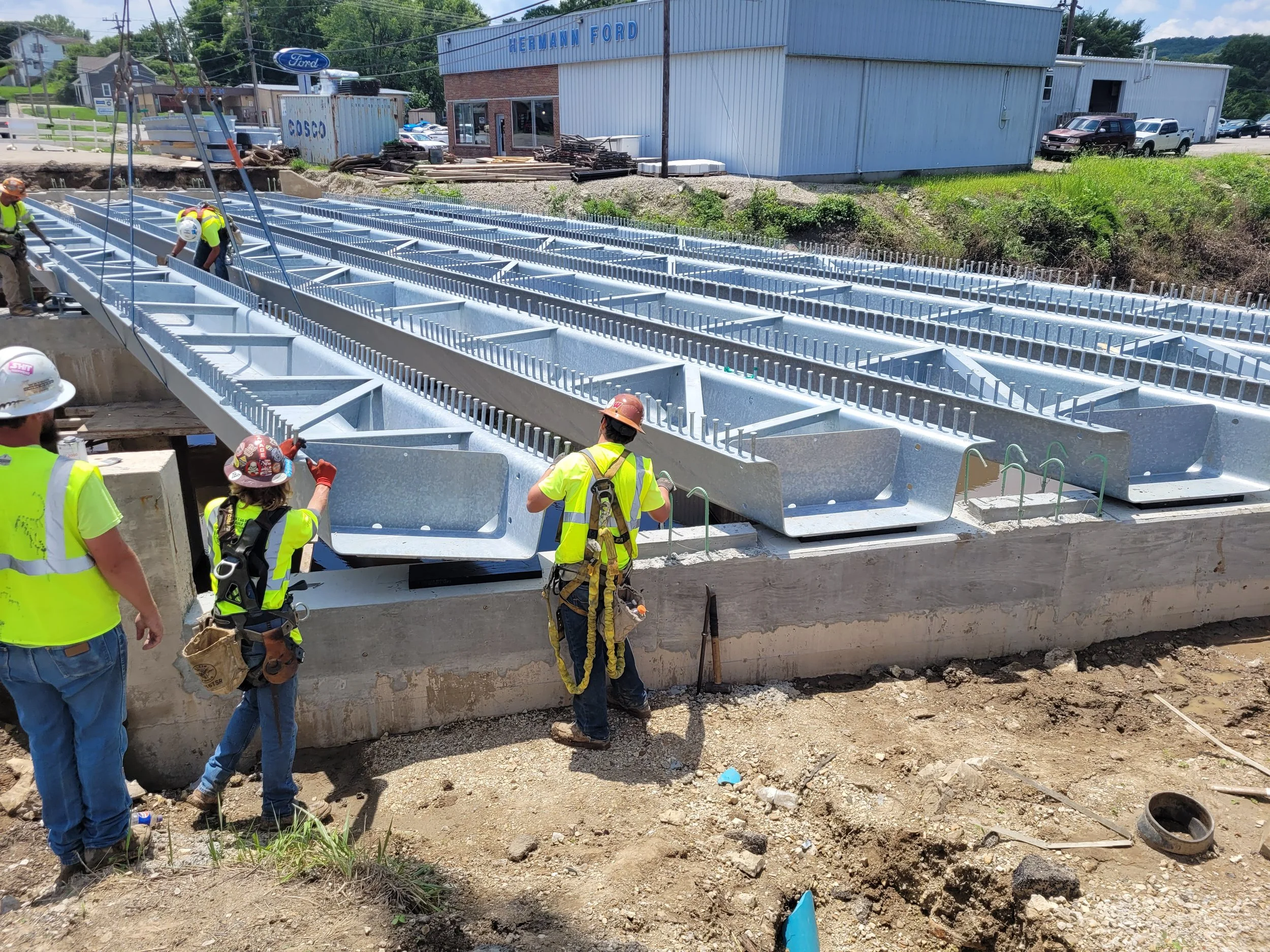Construction workers installing large steel beams at a building site with a Herrmann Ford dealership and parked cars in the background.