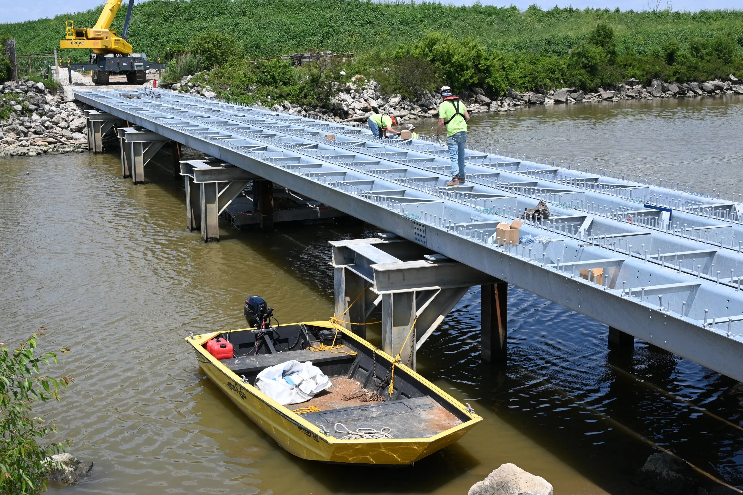 Construction workers installing a metal bridge over a river, with a yellow boat in the foreground, a yellow crane in the background, and lush green trees along the riverbank.