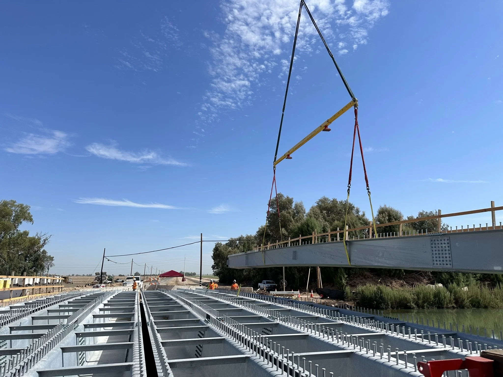 Construction workers are installing a large metal bridge over a body of water under a blue sky with scattered clouds.