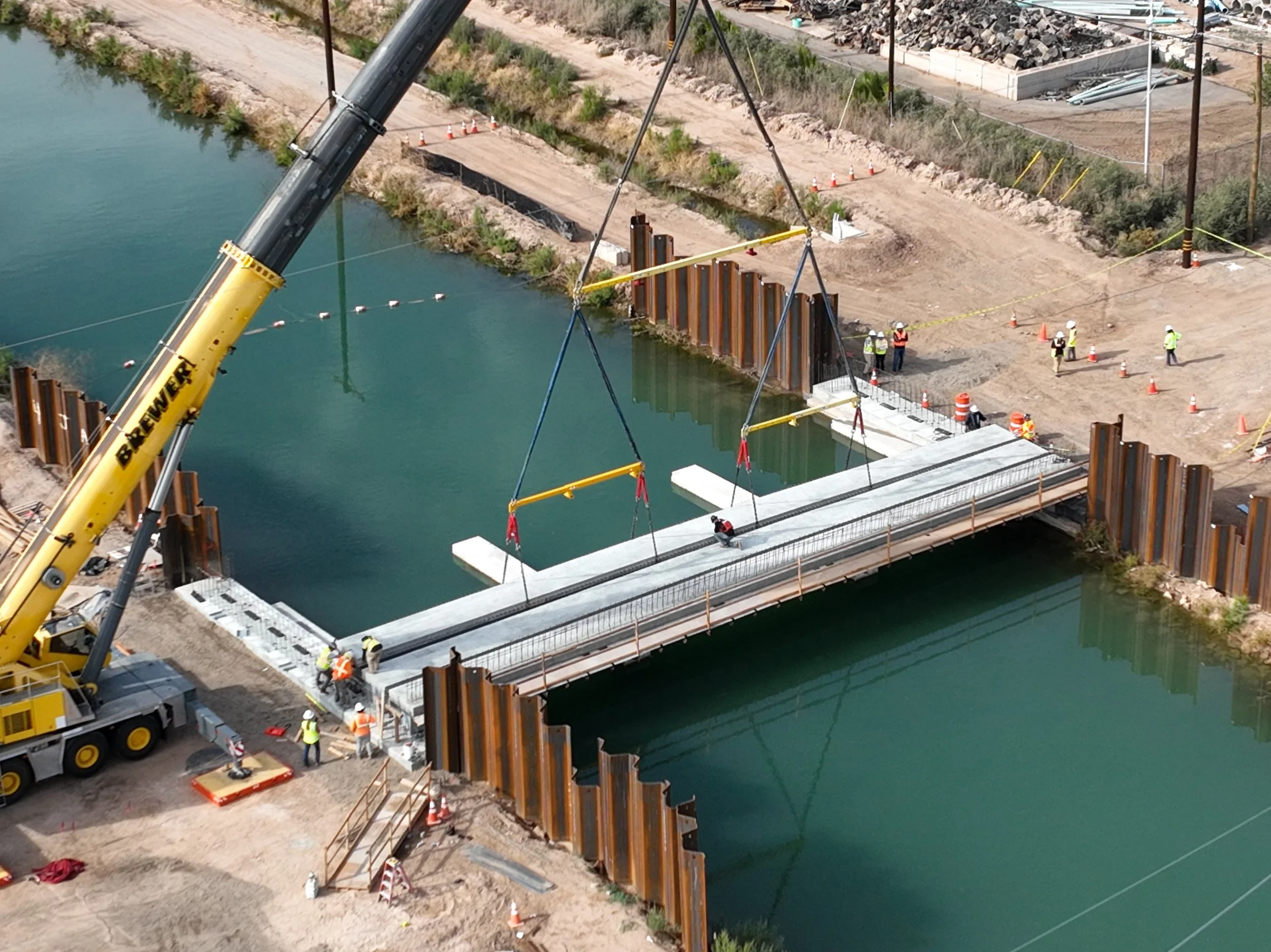 Construction workers and a crane building a bridge over a body of water, with steel and concrete structures and a nearby dirt road.