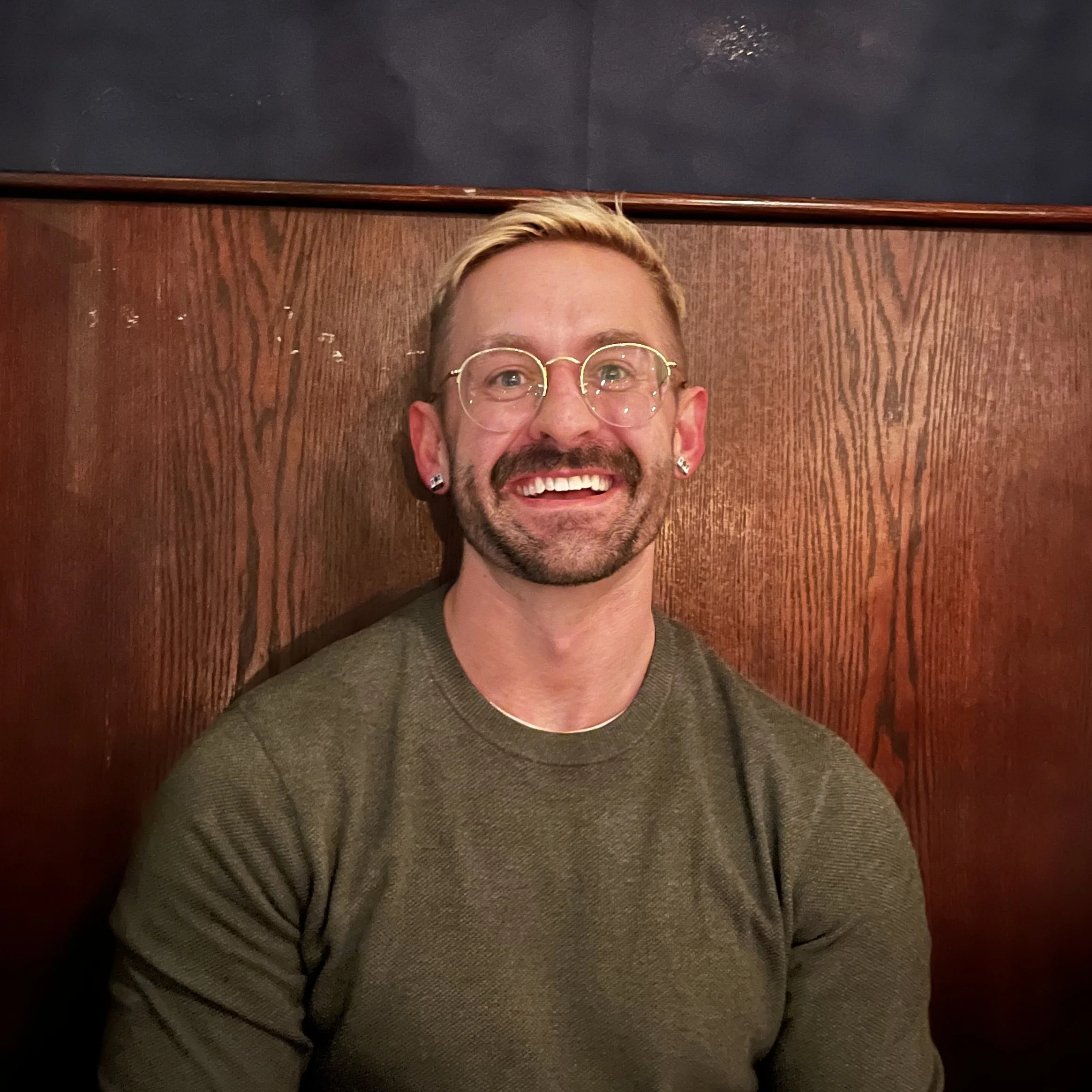 A smiling man with glasses, earrings, and a beard sits against a wooden wall.