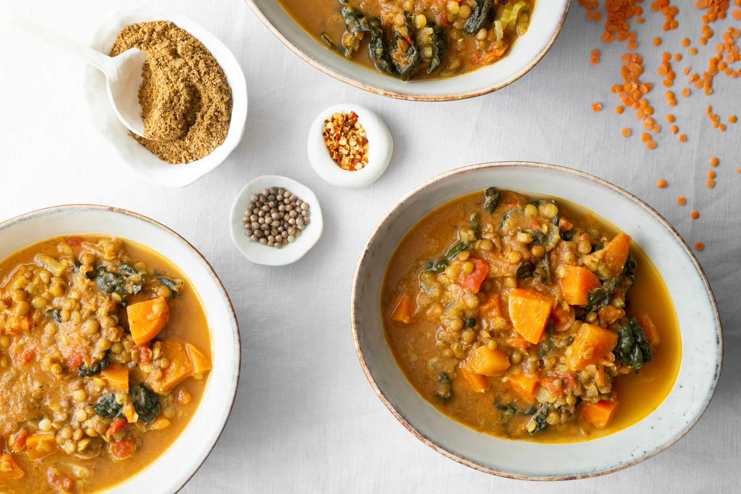 Bowl of lentil and vegetable soup with carrots, spinach, and tomatoes, served in ceramic bowls on a white tablecloth, with small bowls of spices and chili flakes nearby.