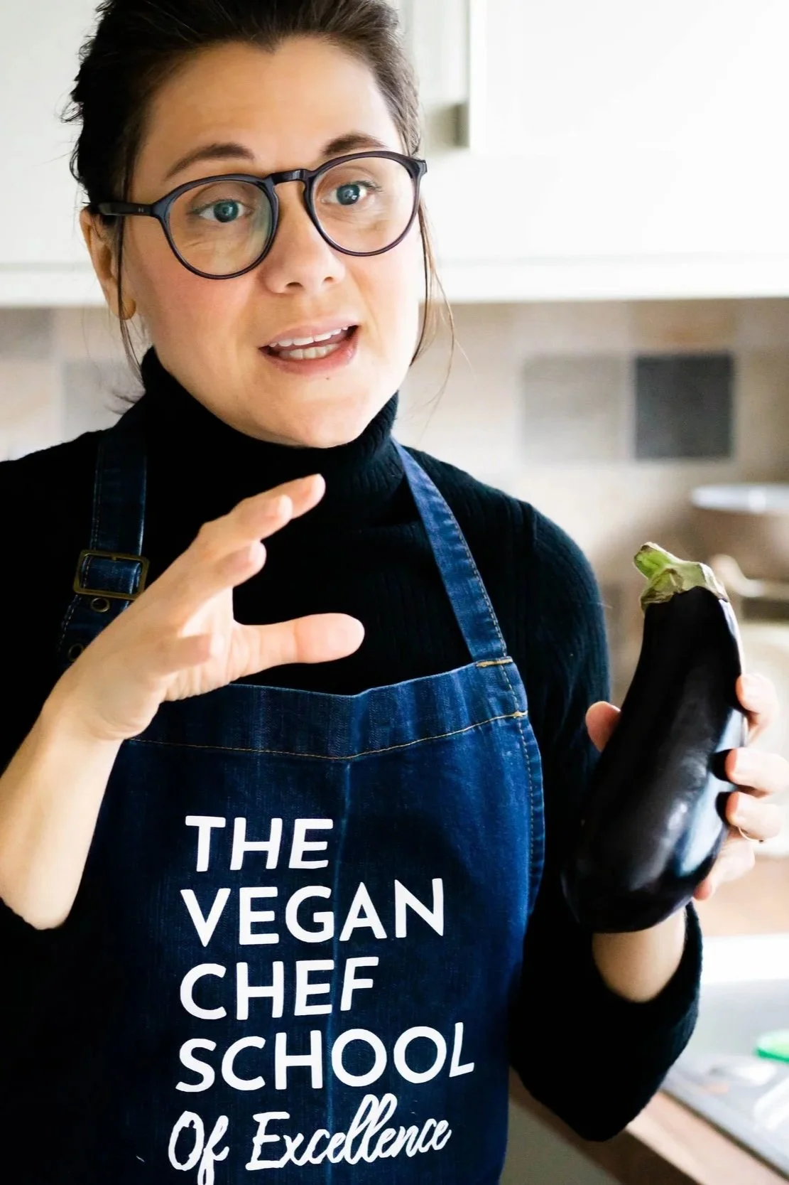 A woman wearing glasses and a black turtleneck, holding an eggplant in her right hand, and wearing a blue apron that says 'The Vegan Chef School of Excellence'.