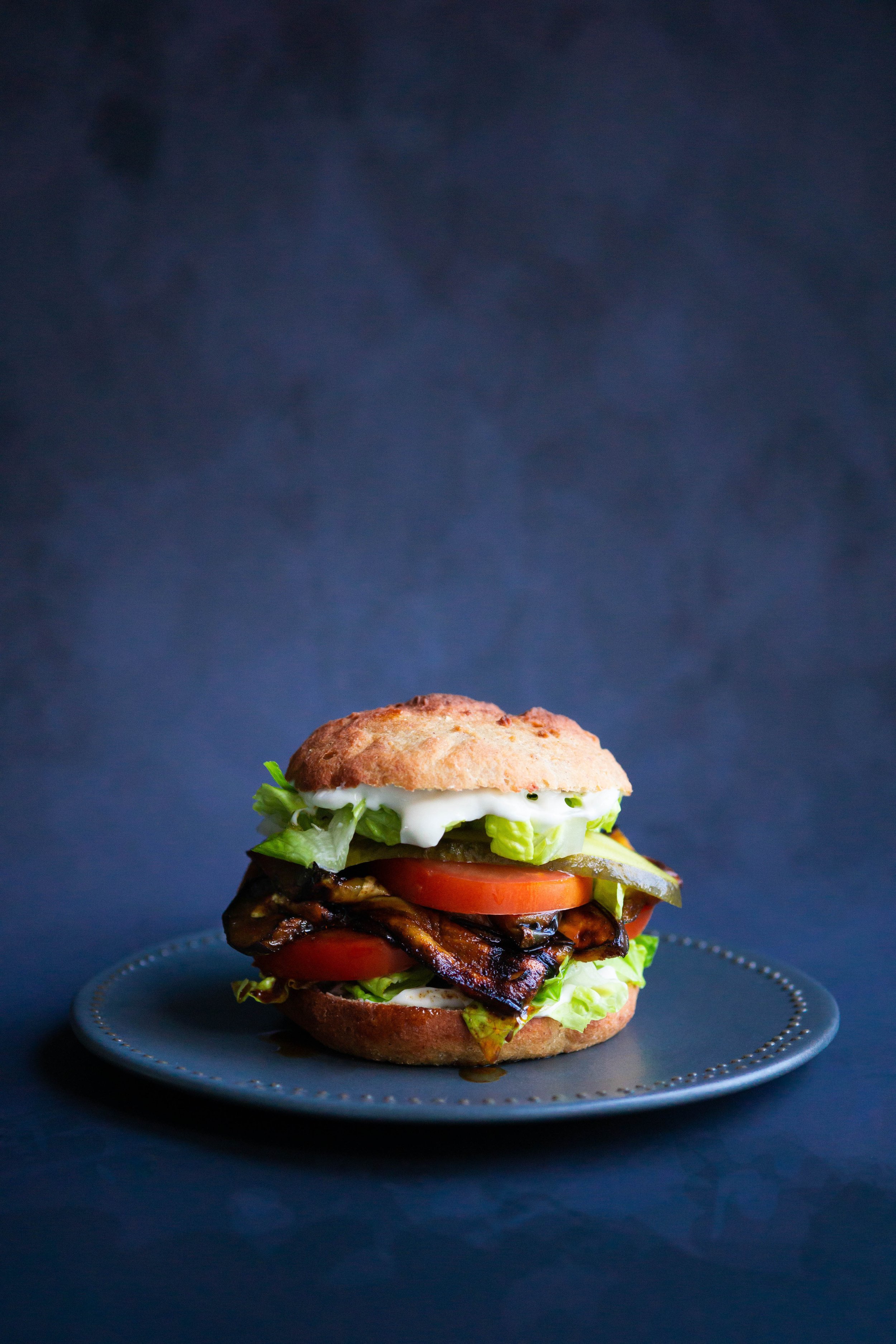 A gourmet sandwich on a gray plate, filled with lettuce, tomato, smoked meat, pickles, and topped with mayonnaise or dressing, against a dark background.