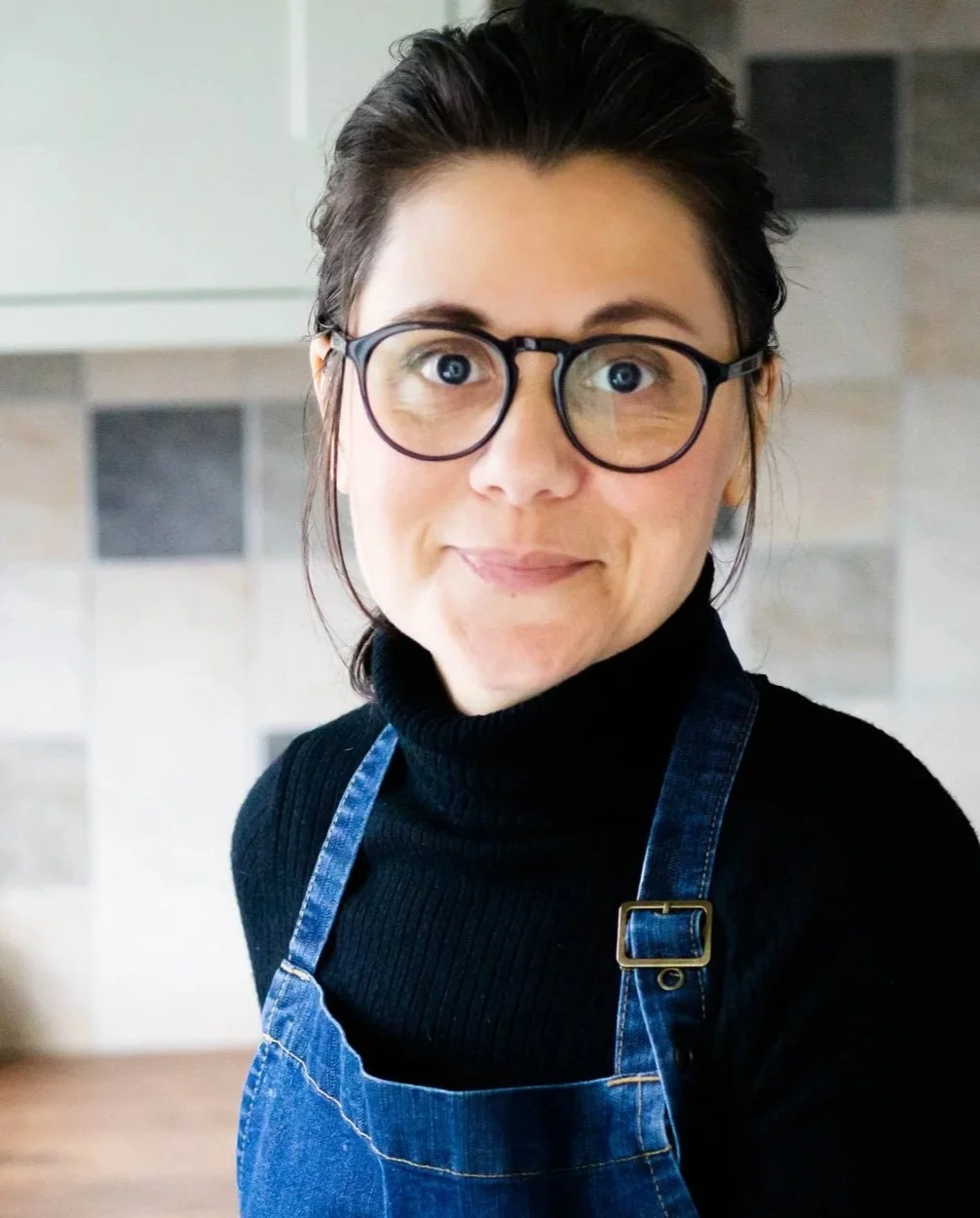 A woman wearing glasses, a black turtleneck, and a denim apron, standing in a kitchen with tiled walls.