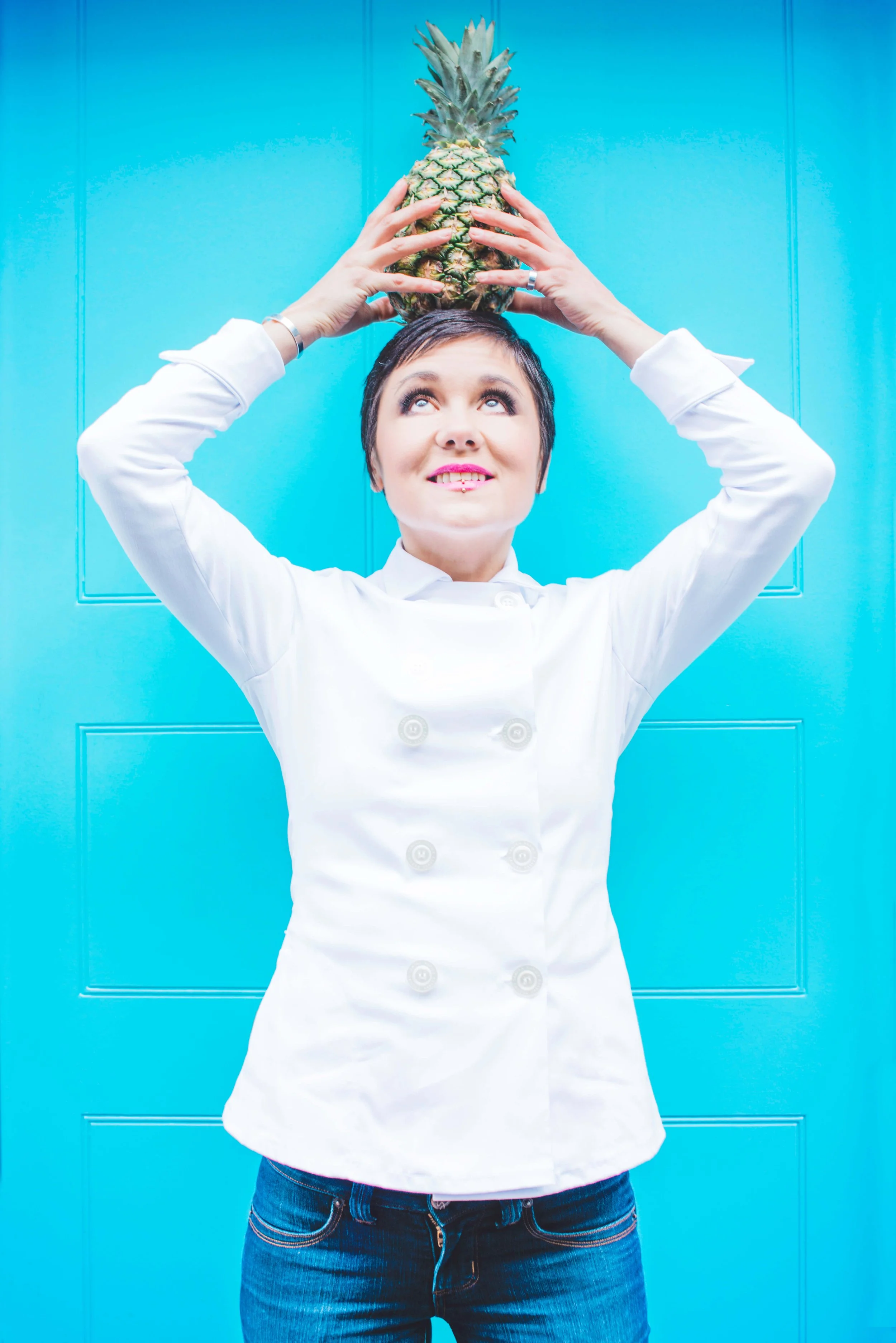 Woman with short dark hair and bright lipstick holding a pineapple above her head in front of a vibrant blue background.