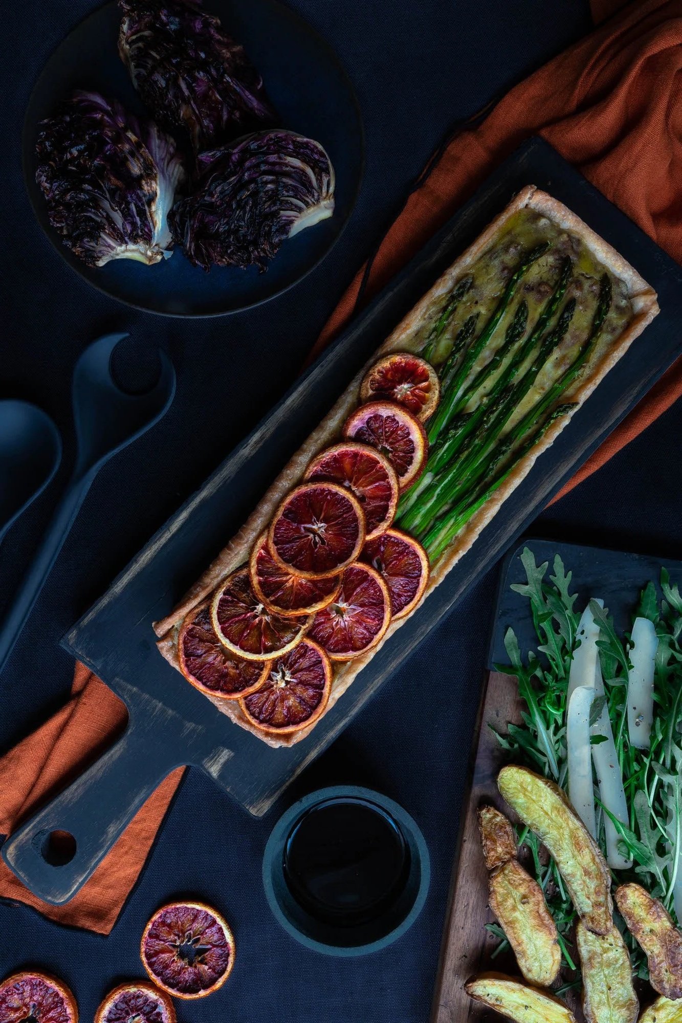 A flat lay of a rectangular wooden serving board with a salmon en croûte topped with blood orange slices and green asparagus, surrounded by roasted sweet potato wedges, arugula salad, a small bowl of red wine, and purple endive on dark-colored tablecloth.