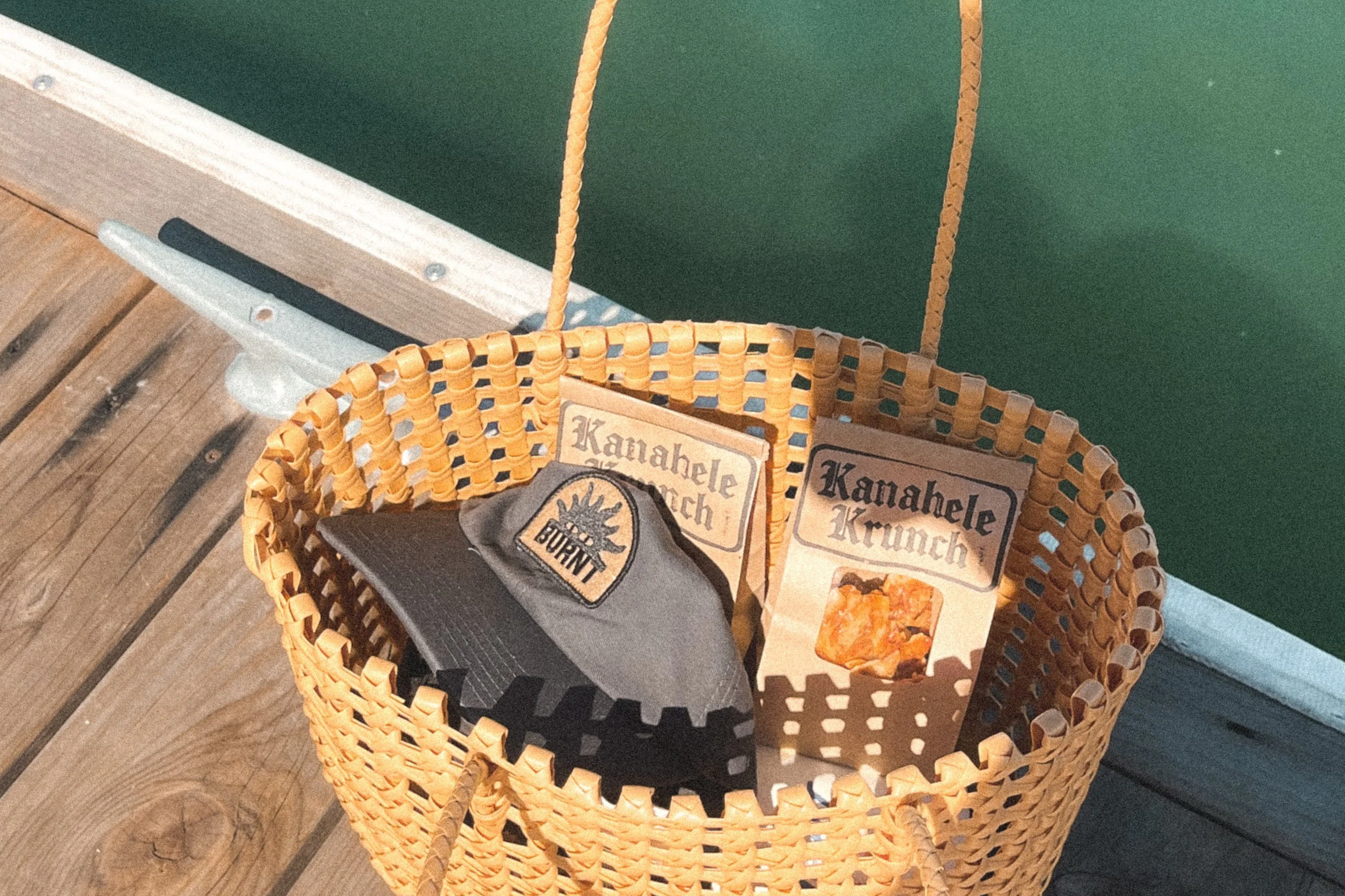 A wicker basket on wooden dock contains a patch, and two booklets labeled "Kahahele Krunch," with the booklet on the right showing an image of crunchy snack.