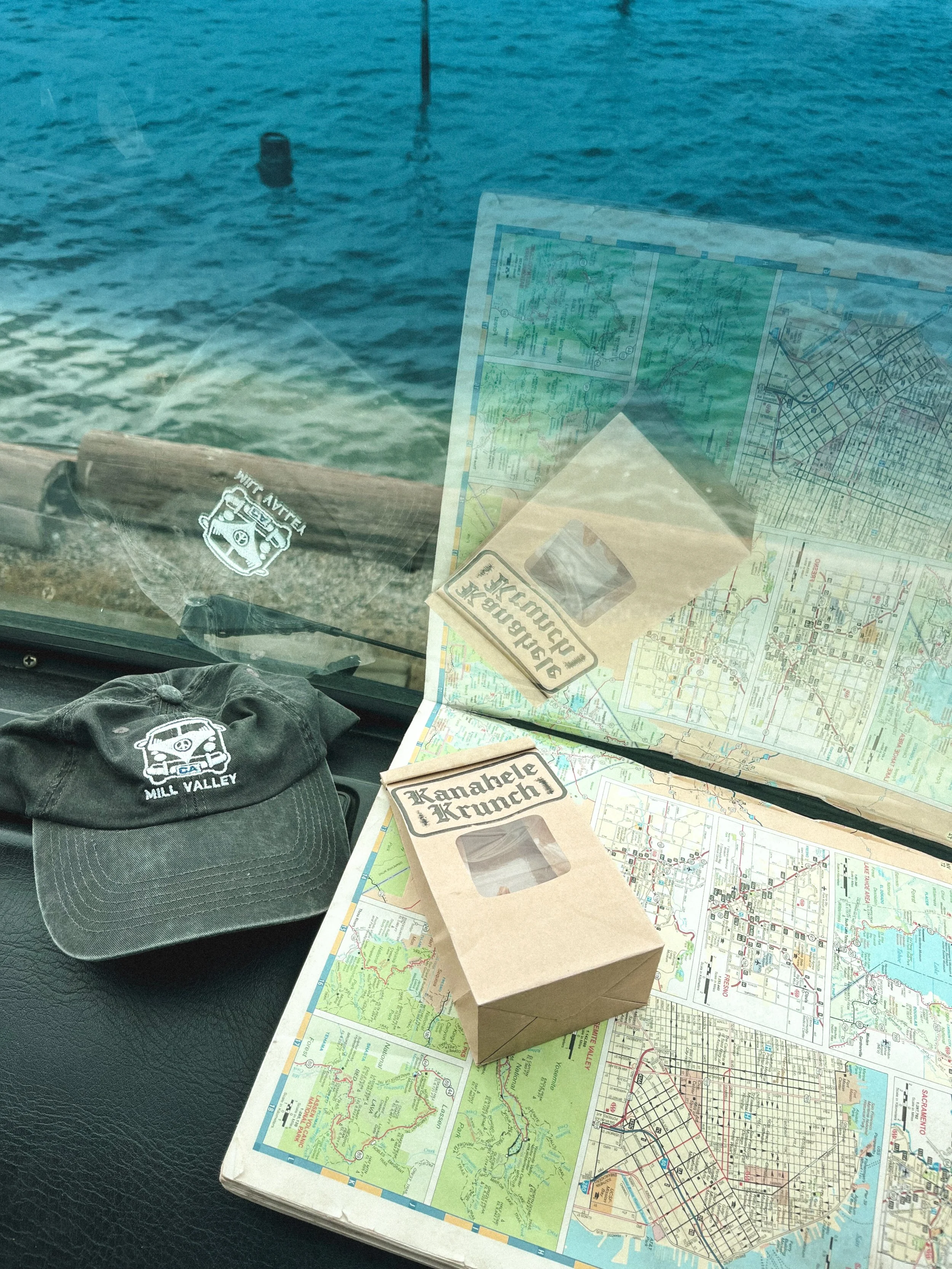 Inside a vehicle, a rolled-up map lays on the dashboard with a black cap that has a logo and the text 'Mill Valley' embroidered on it. Outside, the view of a lake or sea with a buoy and a boat in the distance is visible through the windshield.
