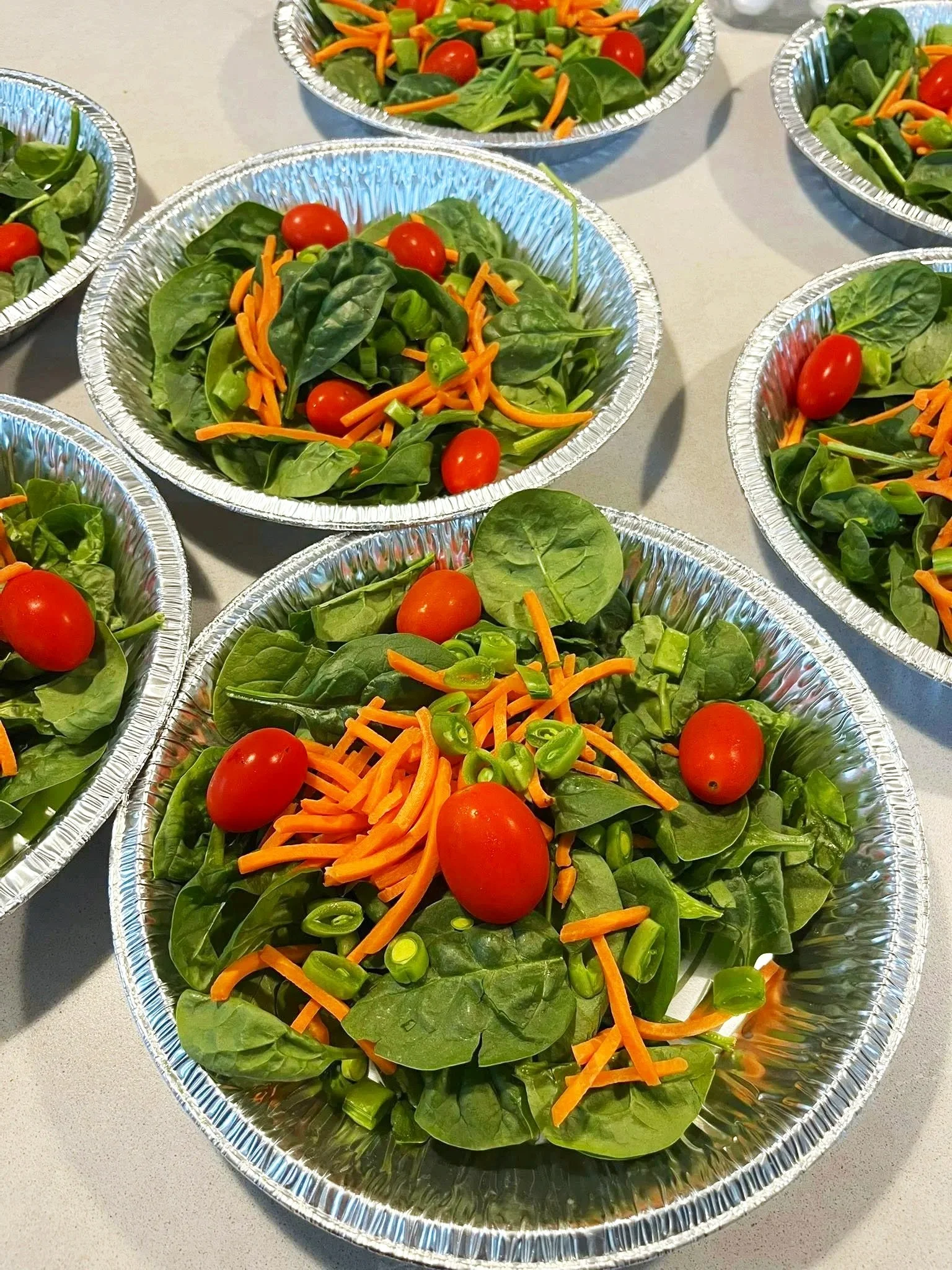 Individual aluminum trays filled with fresh green spinach, cherry tomatoes, shredded carrots, and chopped green onions for a salad buffet.