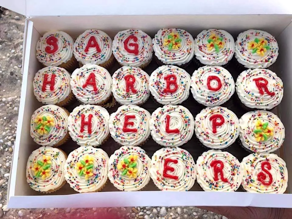 A white box filled with decorated cupcakes spelling out 'SAG HARBOR HELPERS' with colorful sprinkles and floral decorations.