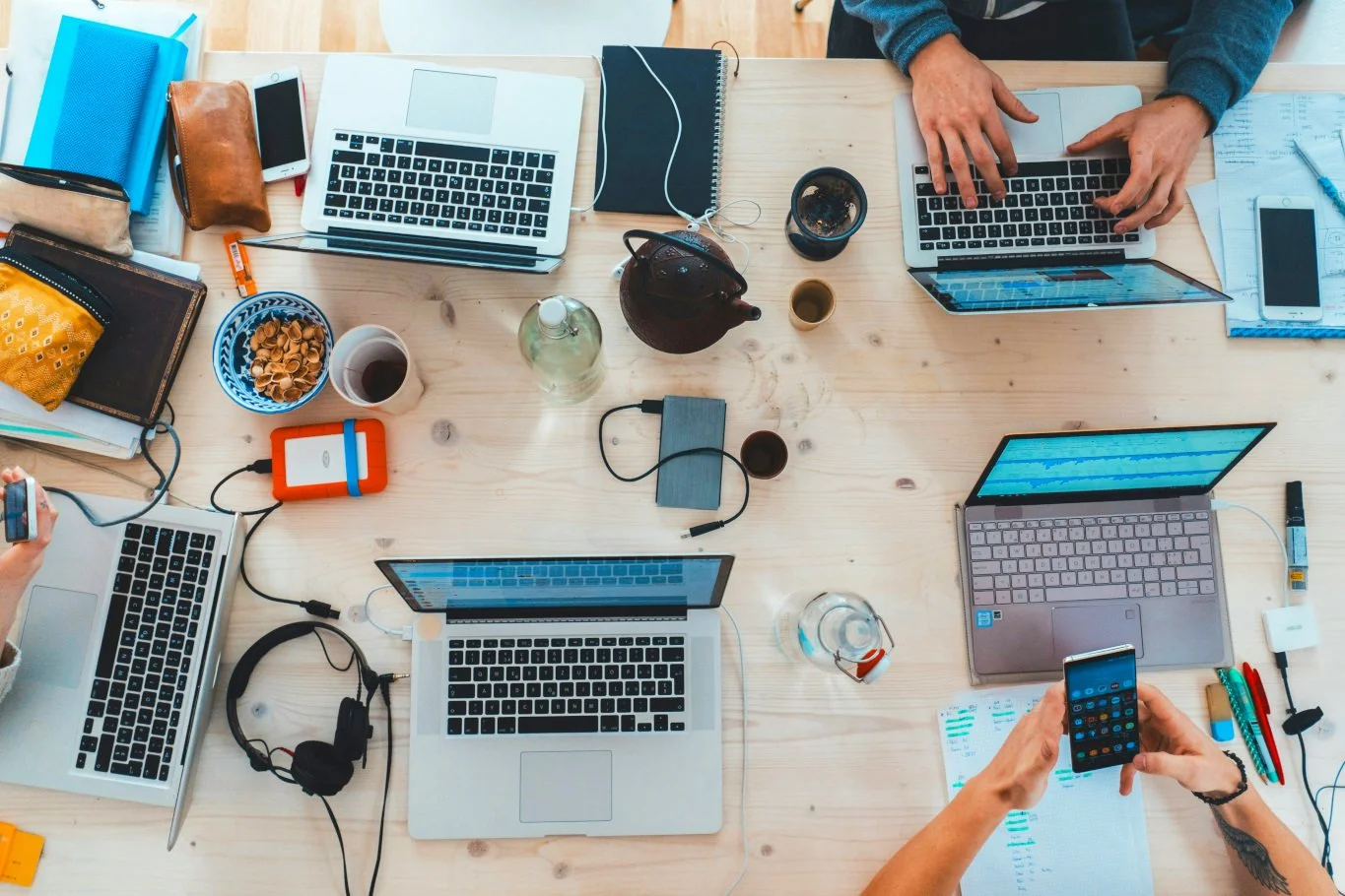 Overhead view of a cluttered wooden table with three people working on laptops, surrounded by notebooks, phones, coffee cups, snacks, and various electronic accessories.