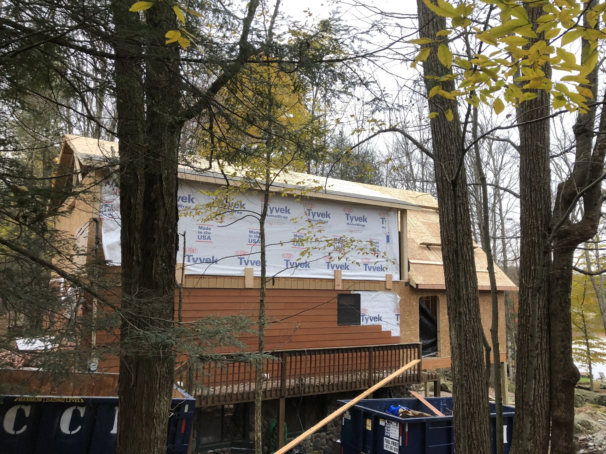 A house under construction in a wooded area with tall trees around it, showing partially finished exterior walls and a roof. Construction materials and debris are visible, including a dumpster.