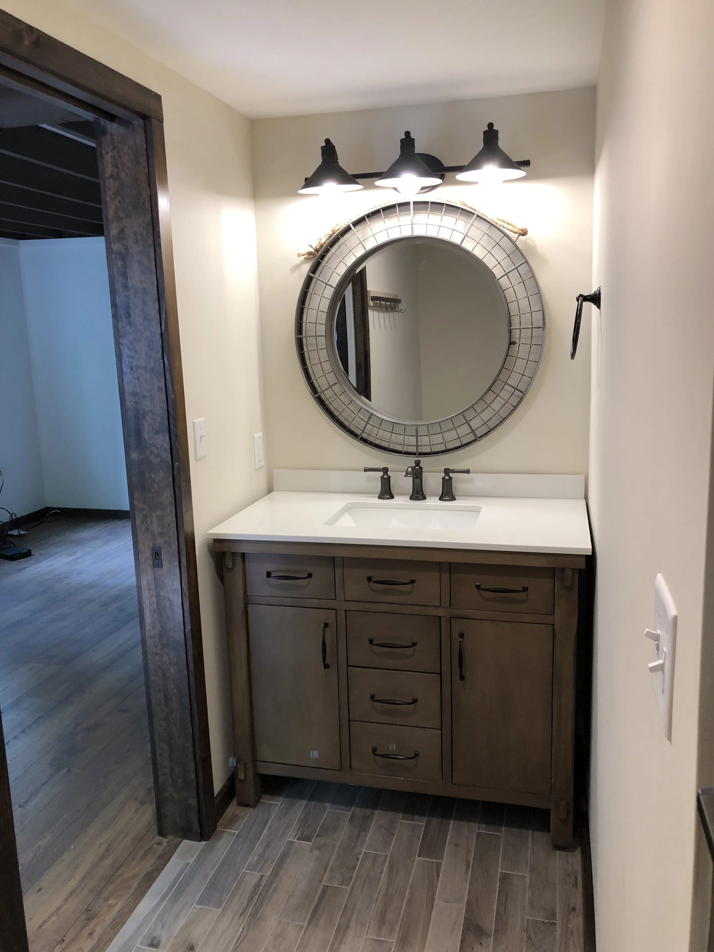 Bathroom vanity with a round mirror, three-bulb light fixture, and wooden cabinet with multiple drawers and handles, located in a room with wooden flooring and beige walls.