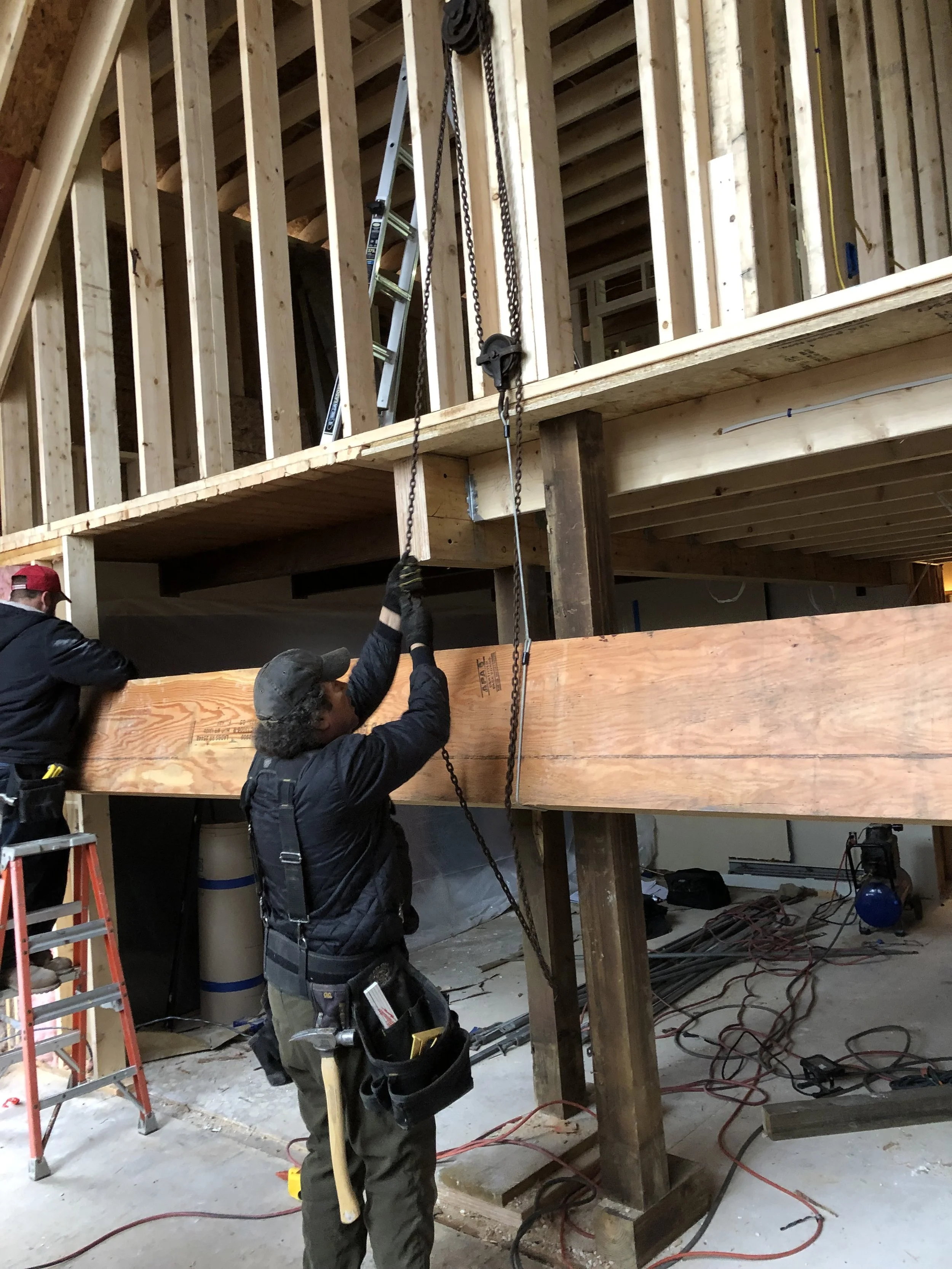 Construction workers installing a wooden beam inside a building under construction, with framing and tools visible.