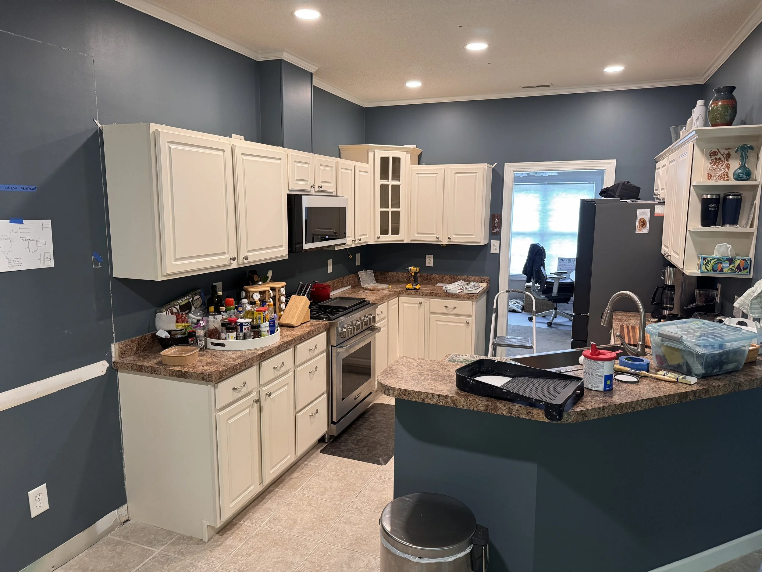 A kitchen with white cabinets, dark blue walls, and granite countertops. There are various kitchen items on the counters, including spices, a cutting board, and cleaning supplies. A window lets in natural light, and a doorway leads to a home office with a chair and window in the background.