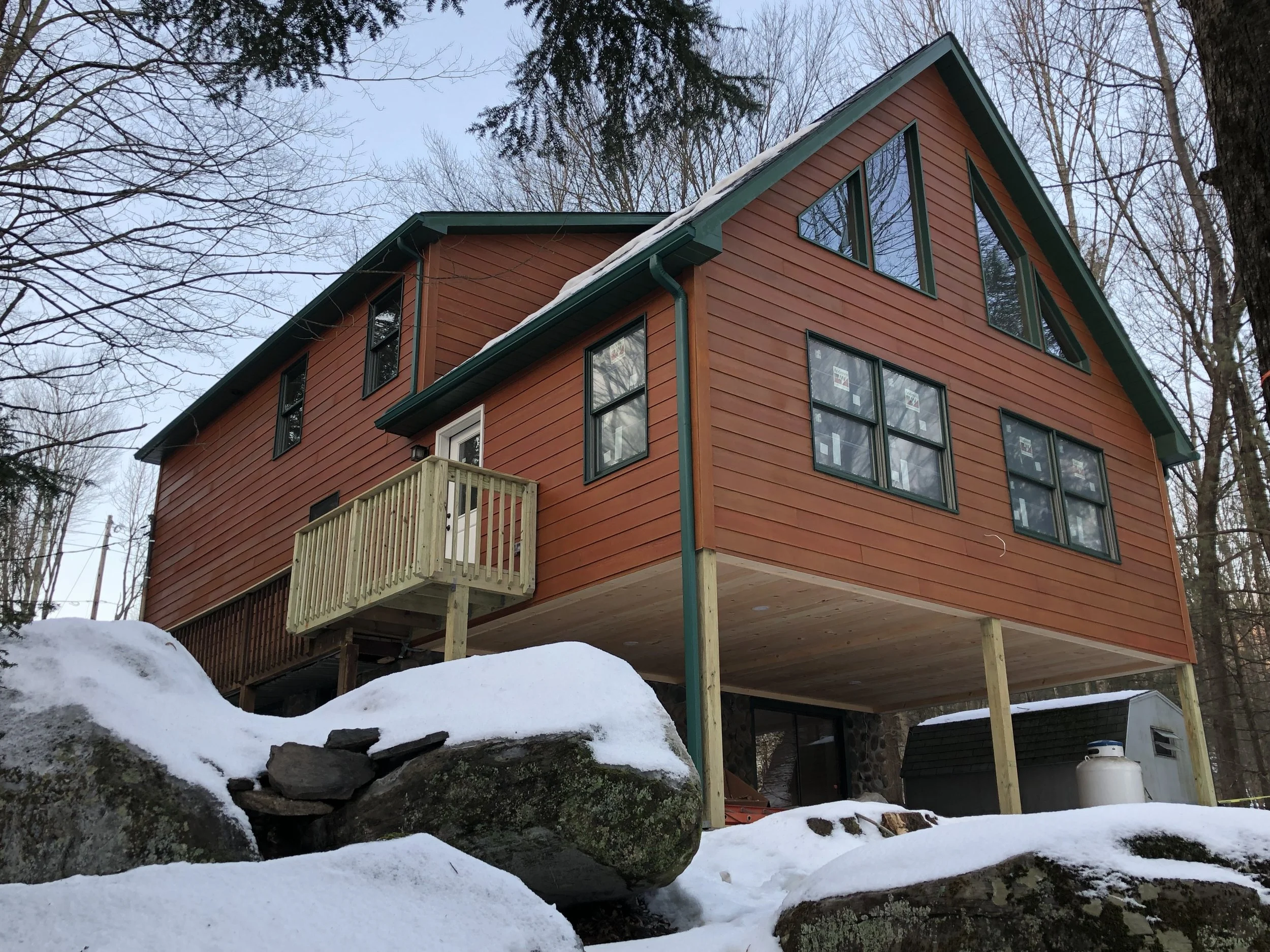 A two-story wooden house with a red exterior and teal trim, raised on stilts with a small balcony and large, triangular windows, set in a snowy wooded area.