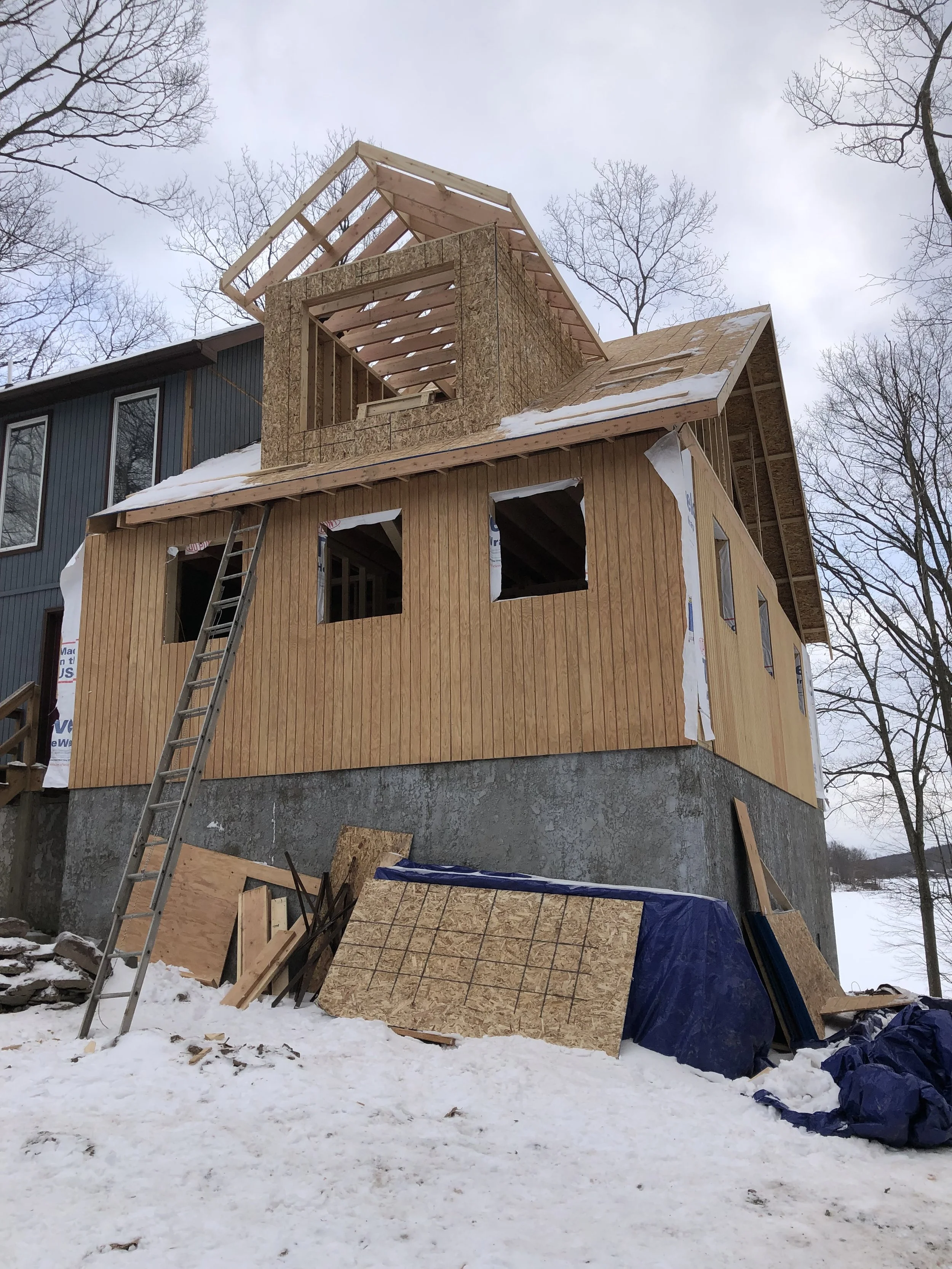 A house under construction with wooden framing and siding, a ladder leaning against it, and construction materials on snow-covered ground, in a winter setting with leafless trees.