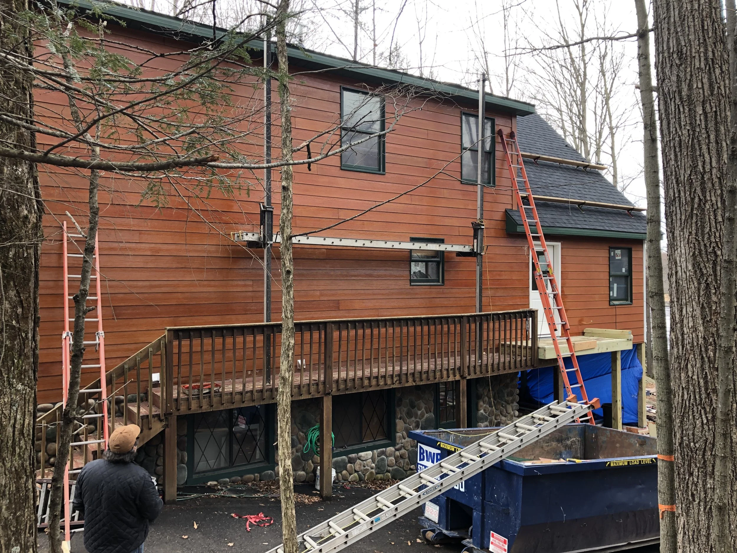 A two-story house under renovation with scaffolding and ladders, surrounded by trees, with a person observing the work.