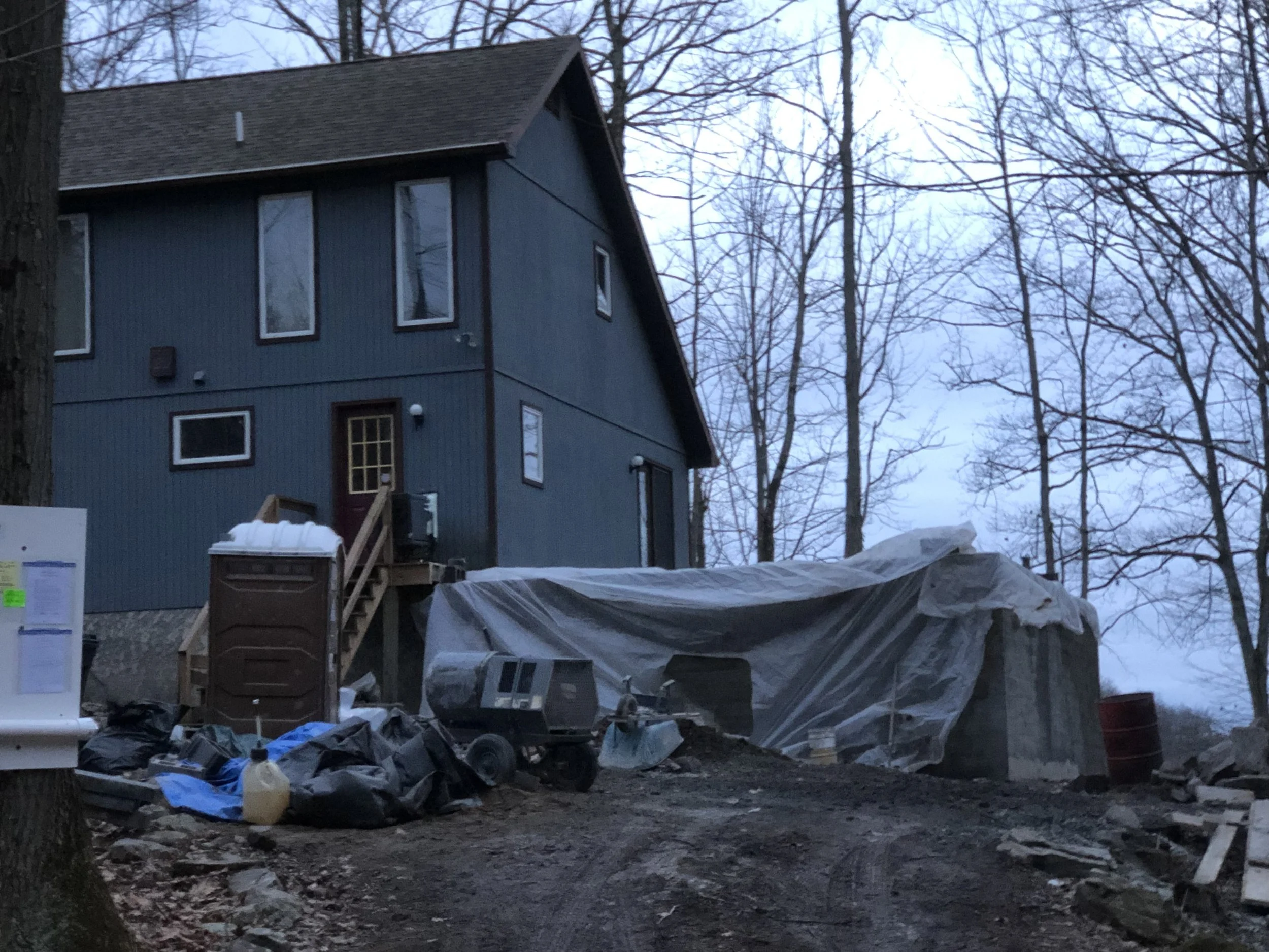 A house under construction or renovation surrounded by construction materials, tools, and debris in a wooded area with leafless trees.