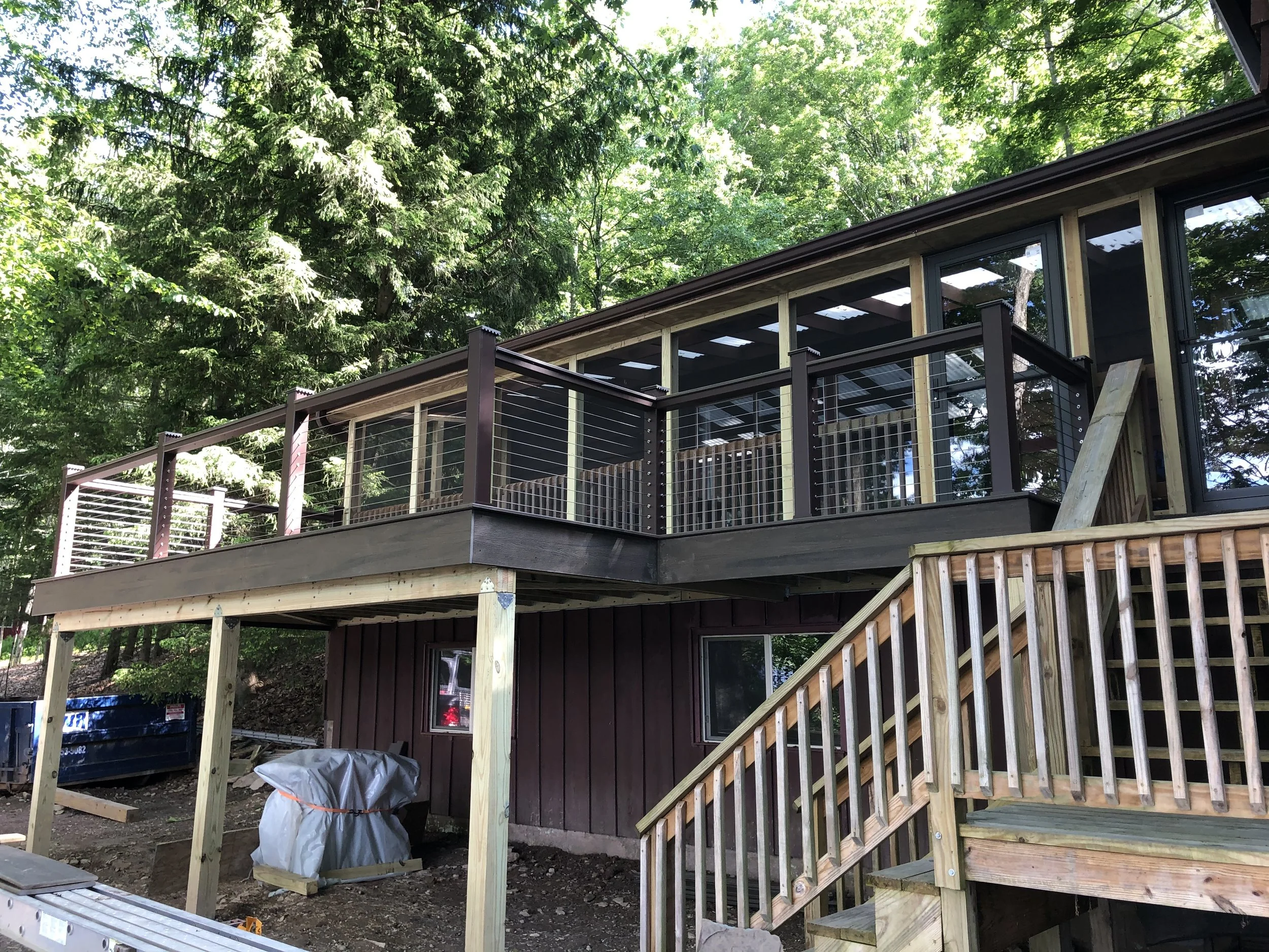 Under construction house with a deck in a wooded area, with stairs leading up to the deck and a staircase going down.