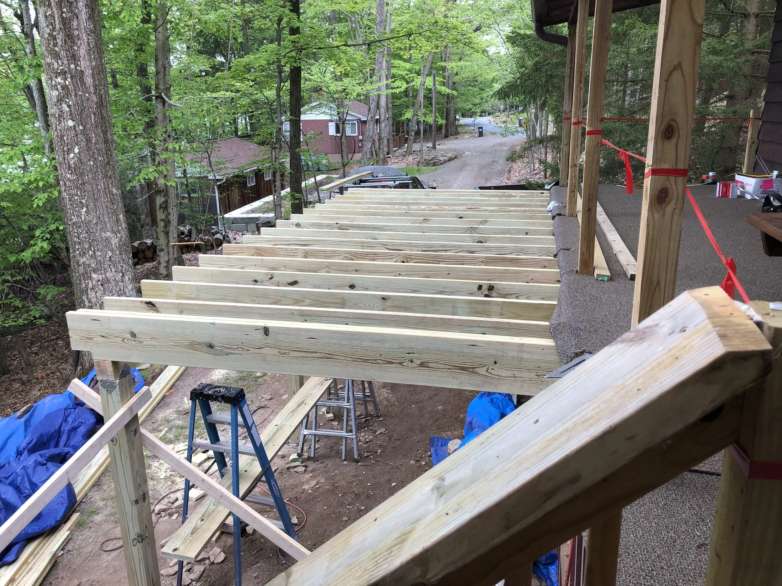 Construction site showing a wooden deck or balcony mostly built with side rails and support beams, surrounded by a forested area with trees and a dirt road.