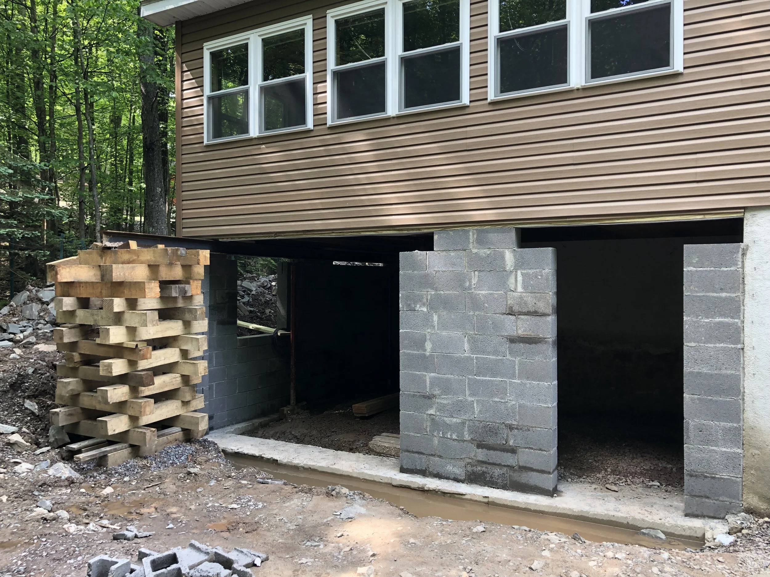 Under-construction house foundation with concrete block walls, with stacked wooden planks on the left. The house is elevated with a brown siding exterior and multiple windows, situated in a wooded area.