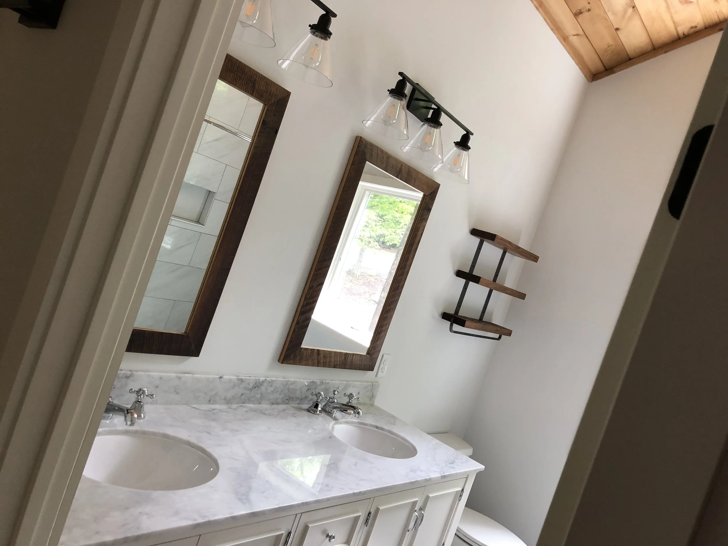 Bathroom with double sink marble countertop, two mirrors with wooden frames, a small window, wall-mounted wooden shelves, and a ceiling light fixture with four glass shades.