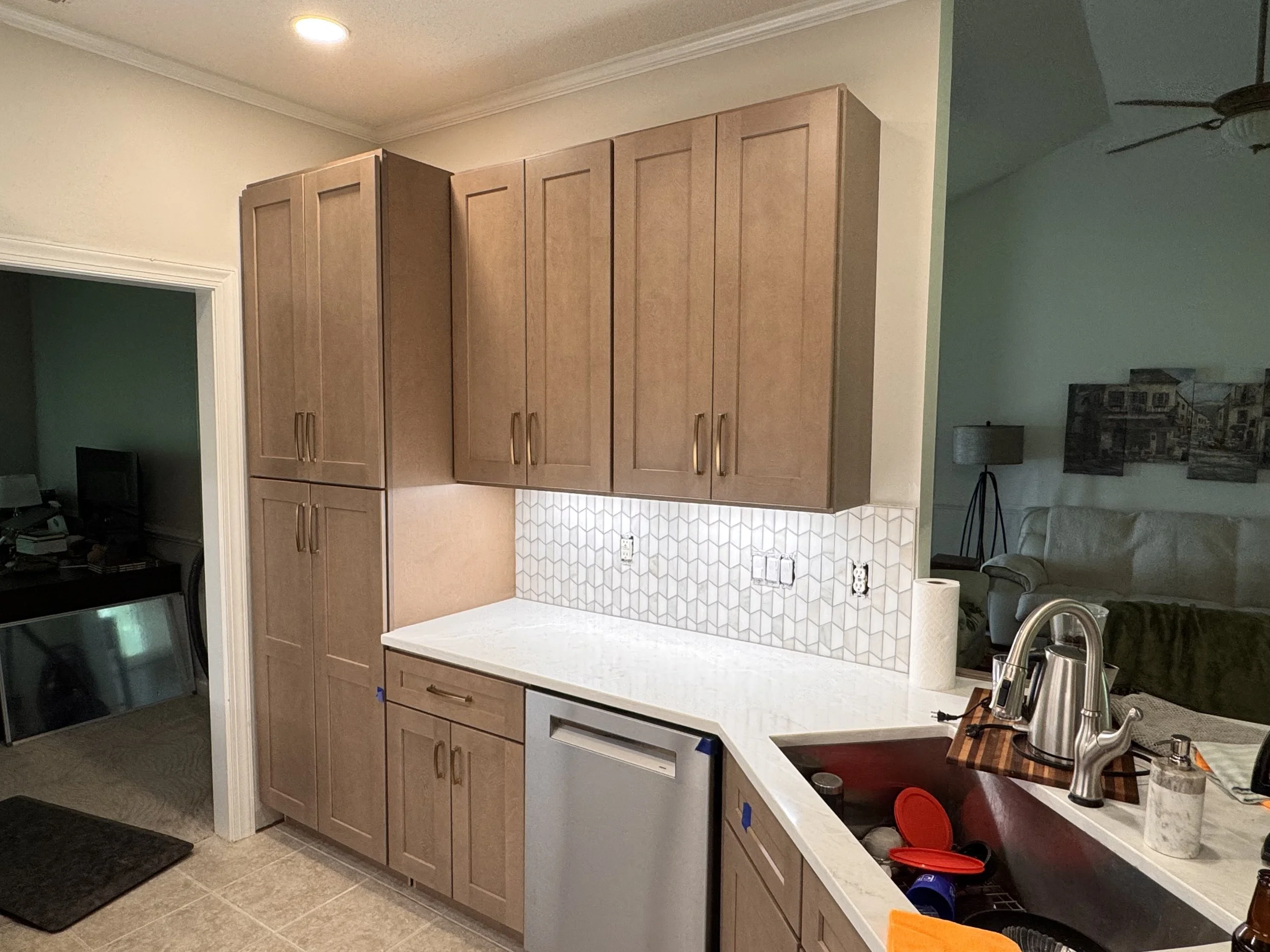 Kitchen with light wooden cabinets, a white countertop, a tile backsplash, a sink with a faucet, and various kitchen items including a paper towel roll, a kettle, and containers. Part of a living room with a sofa and artwork is visible in the background.