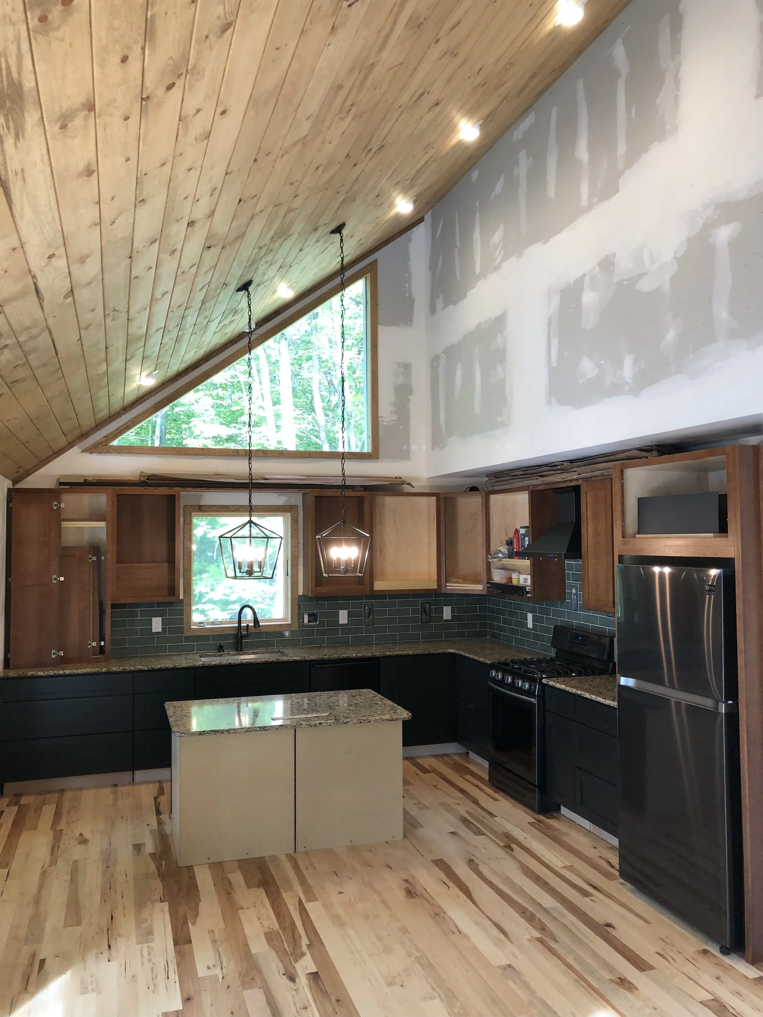 Kitchen with dark green lower cabinets, open wooden upper cabinets, granite countertops, a small island, black appliances, and a large triangular window with a view of trees, under a wood-paneled ceiling with recessed lighting.