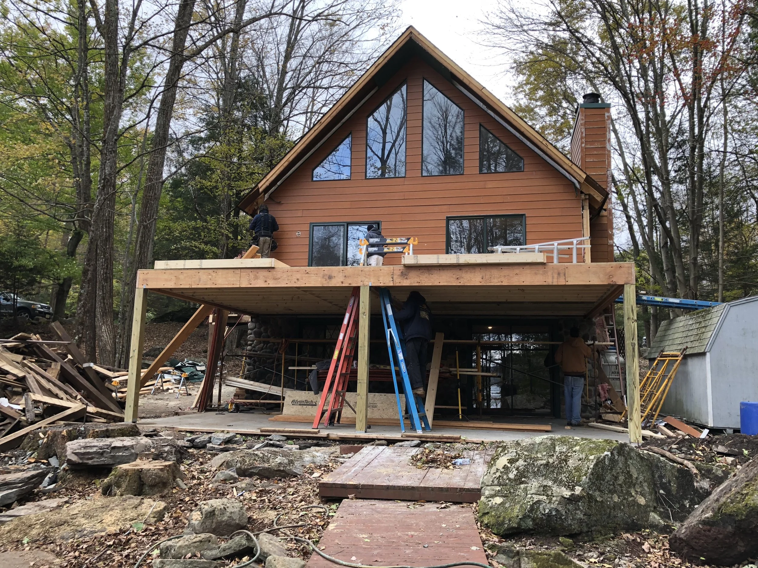 A two-story house under construction with a wooden deck on the second floor. There are workers on the deck and standing underneath, with construction equipment and materials scattered around. The house is surrounded by trees and large rocks.