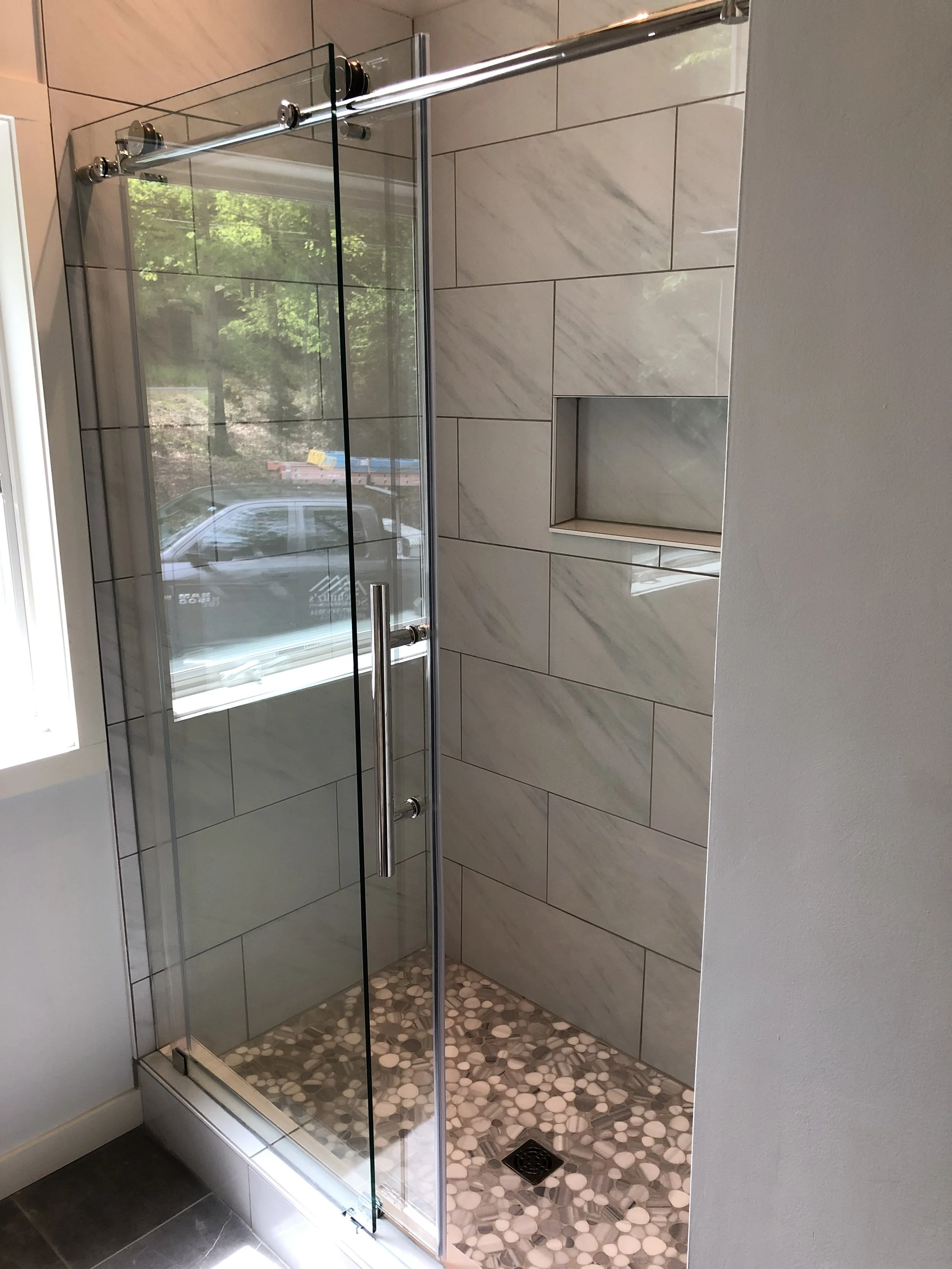 A modern glass shower with a sliding door, tiled walls, a built-in niche, and pebble stone flooring, reflecting a window with greenery outside.