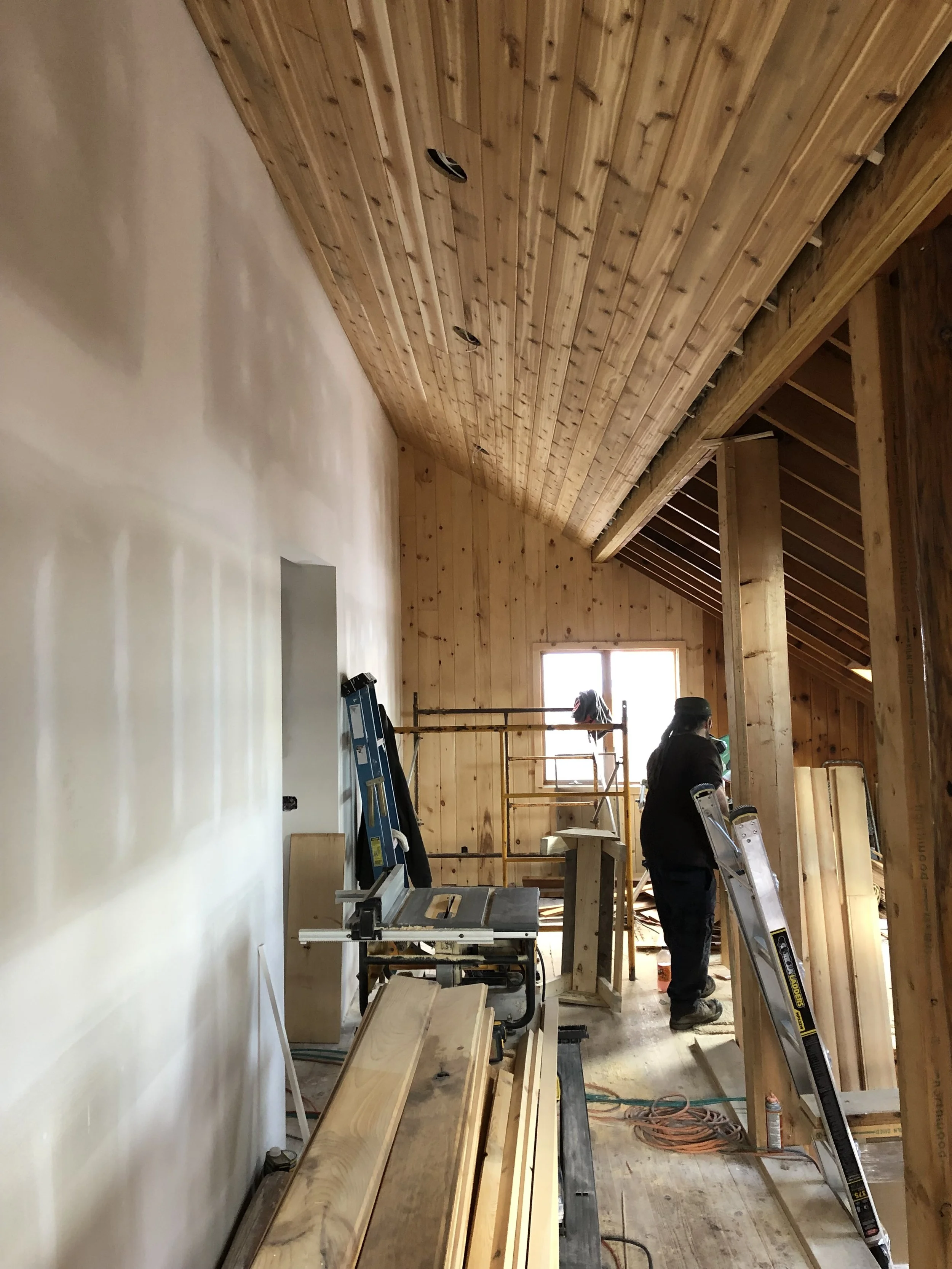 Interior view of a room under construction with wood paneling on ceiling and walls, tools and construction materials scattered, and workers installing or working on the carpentry.